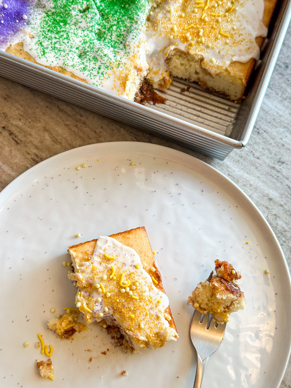 A white plate with a slice of king cake with the cake pan in the background with the king cake inspired sheet cake. 