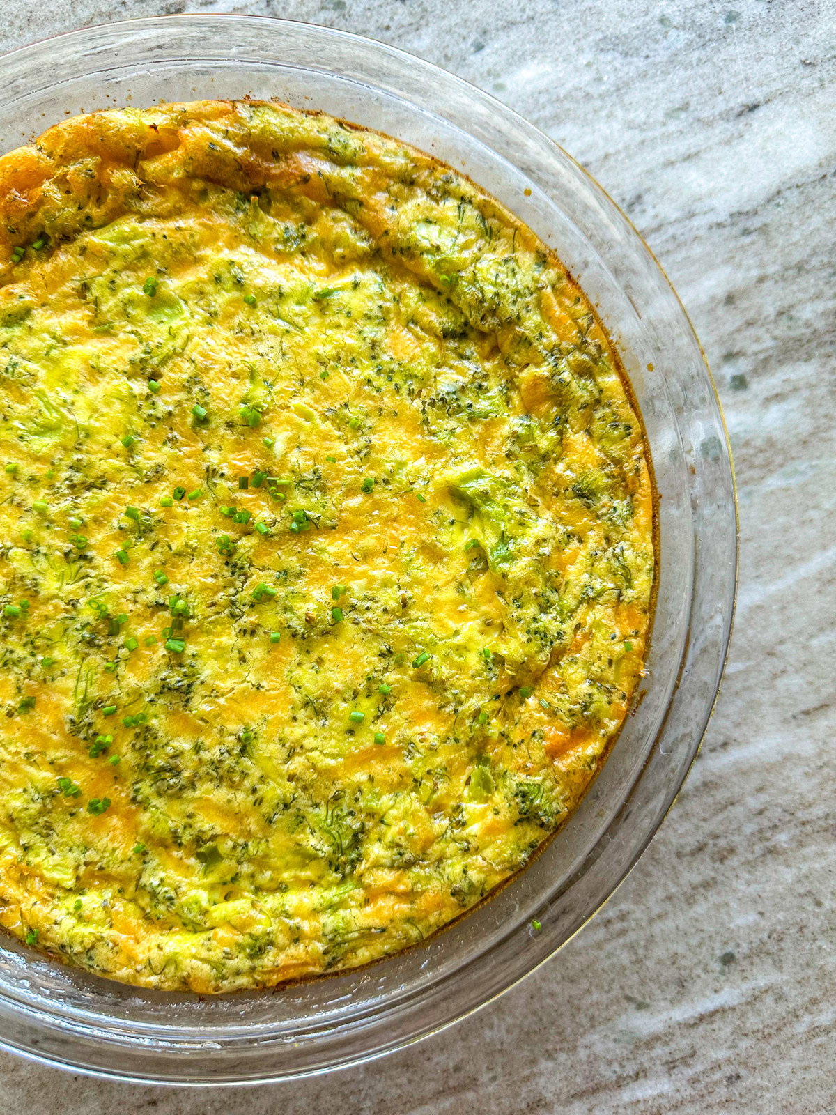 A broccoli and cheese quiche topped with chives in a glass pie dish on the countertop. 