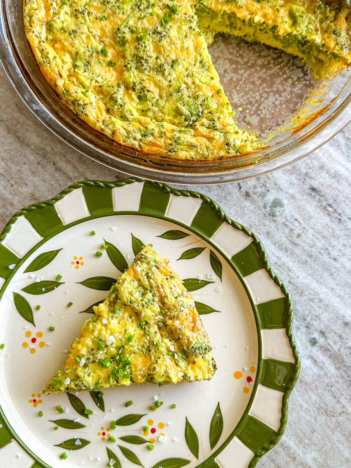 Slice of broccoli and cheese quiche on a floral green plate with a pie dish of quiche in the background.