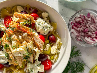A white bowl filled with tzatziki sauce, pasta, chicken, cherry tomatoes, feta, chopped dill, pepperoncinis, Kalamata olives, marinated artichoke hearts with a bowl of feta and a sprig of dill in the background.