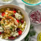 A white bowl filled with tzatziki sauce, pasta, chicken, cherry tomatoes, feta, chopped dill, pepperoncinis, Kalamata olives, marinated artichoke hearts with a bowl of feta and a sprig of dill in the background.