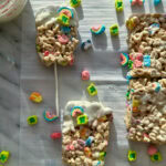 A white marble board with lucky charms bars on top dipped in white chocolate and sprinkled with lucky charms marshmallows.