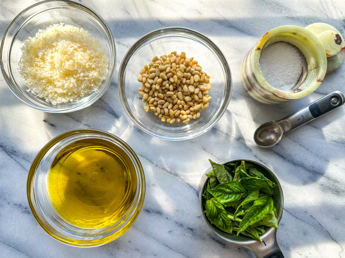 Clear ingredients bowls filled with parmigiano reggiano, pine nuts, basil, olive oil, and kosher salt. 