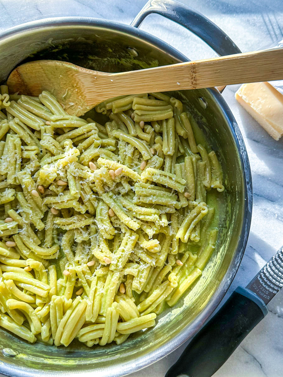 A saucepan of pasta in a creamy pesto sauce with a wooden spoon, block of cheese, and grater on the side. 