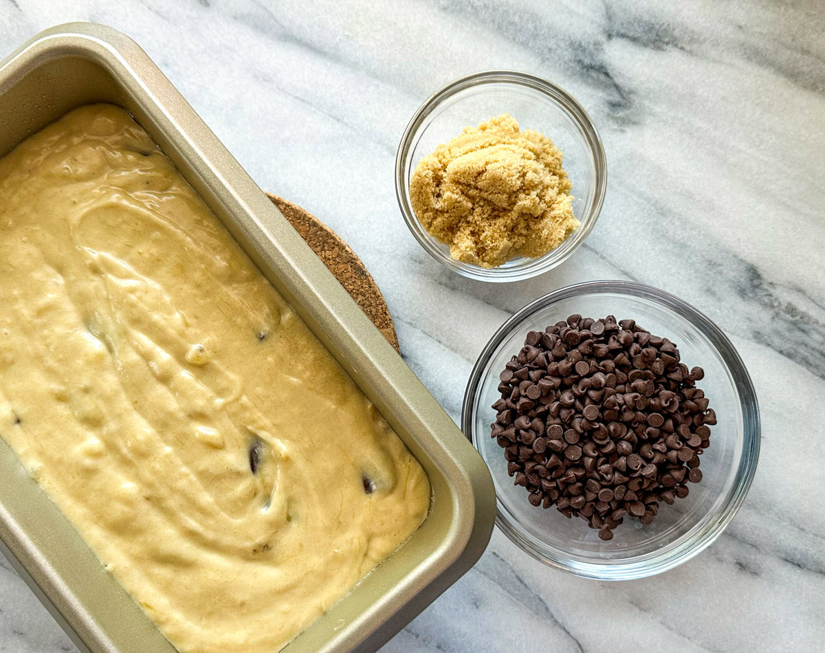 Uncooked banana bread in a loaf pan with a bowl of mini chocolate chips and brown sugar.