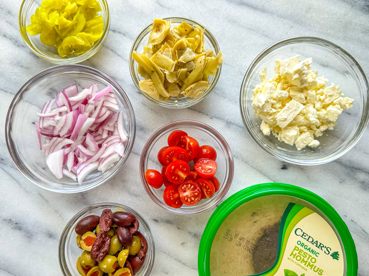 Clear bowls filled with peppers, marinated artichoke hearts, feta, chopped red onion, cherry tomatoes, green and Kalamata olives and a dish of hummus. 
