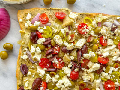 A mediterranean pizza sitting on a marble cutting board topped with hummus, feta, cherry tomatoes, olives, artichoke hearts, and red onion with loose olives in the background.