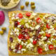 A mediterranean pizza sitting on a marble cutting board topped with hummus, feta, cherry tomatoes, olives, artichoke hearts, and red onion with loose olives in the background.