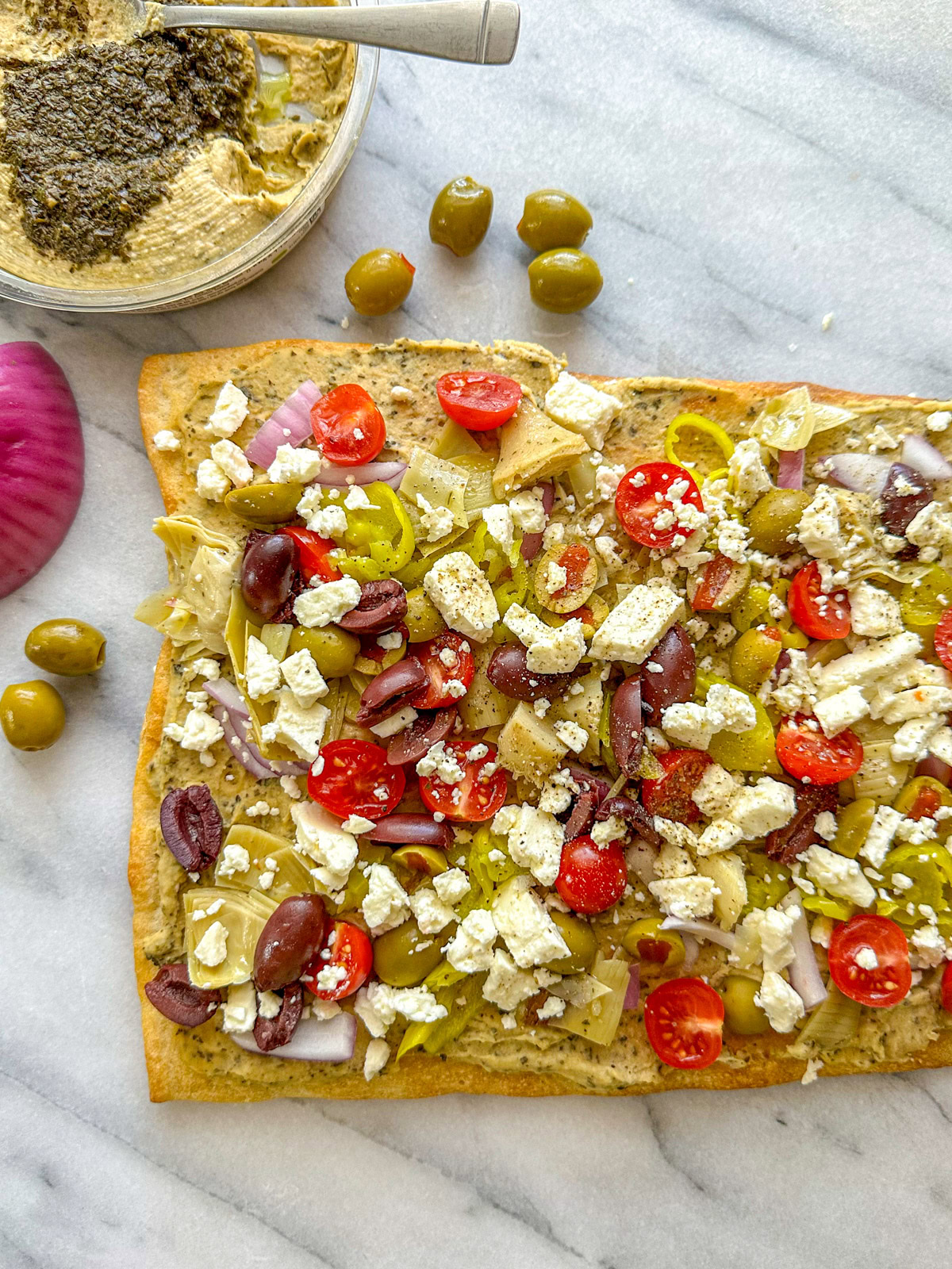 A mediterranean pizza sitting on a marble cutting board topped with hummus, feta, cherry tomatoes, olives, artichoke hearts, and red onion with loose olives in the background.