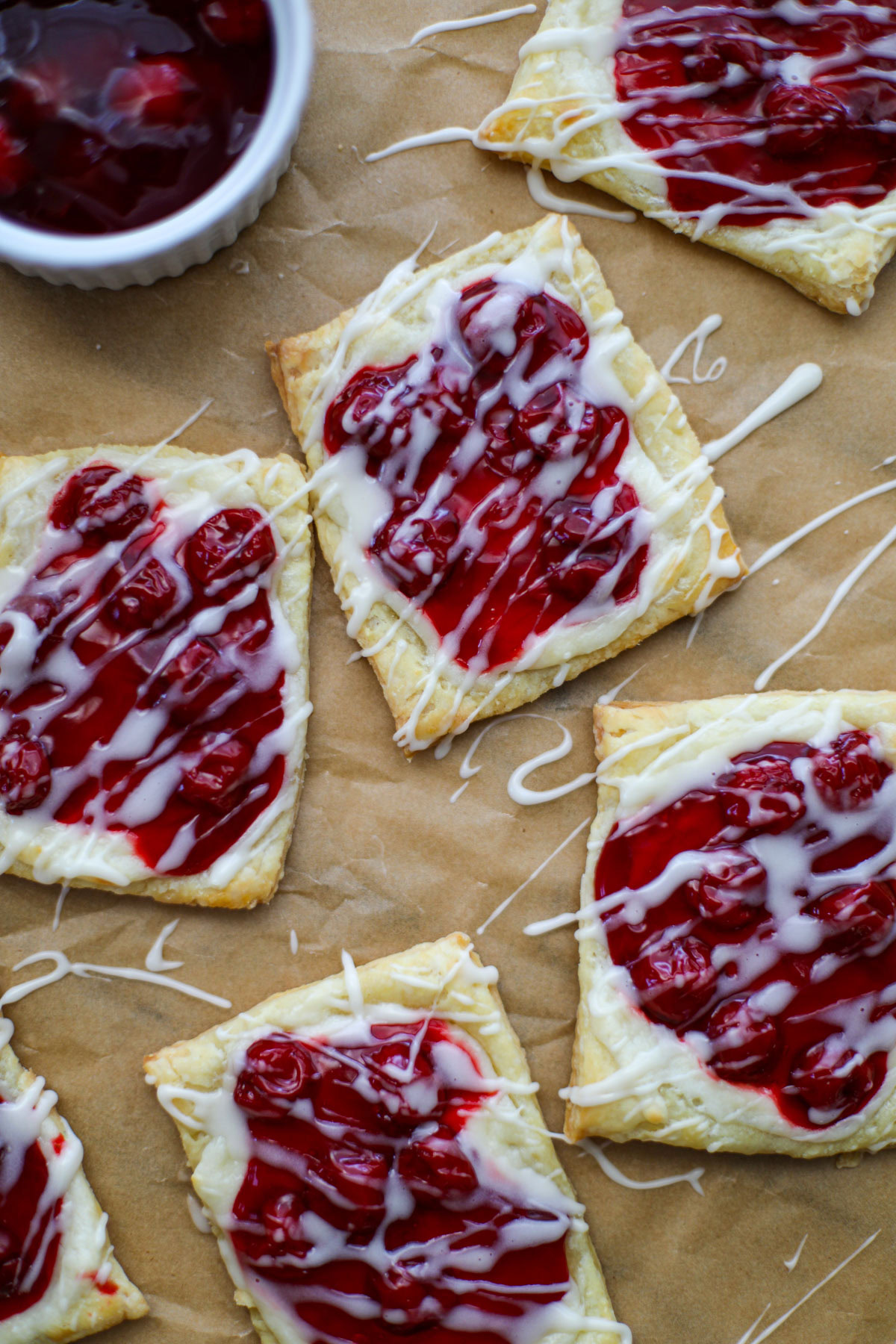 Brown parchment paper topped with square cherry cheese danishes drizzled with a white glaze with a bowl of cherry pie filling in the background.