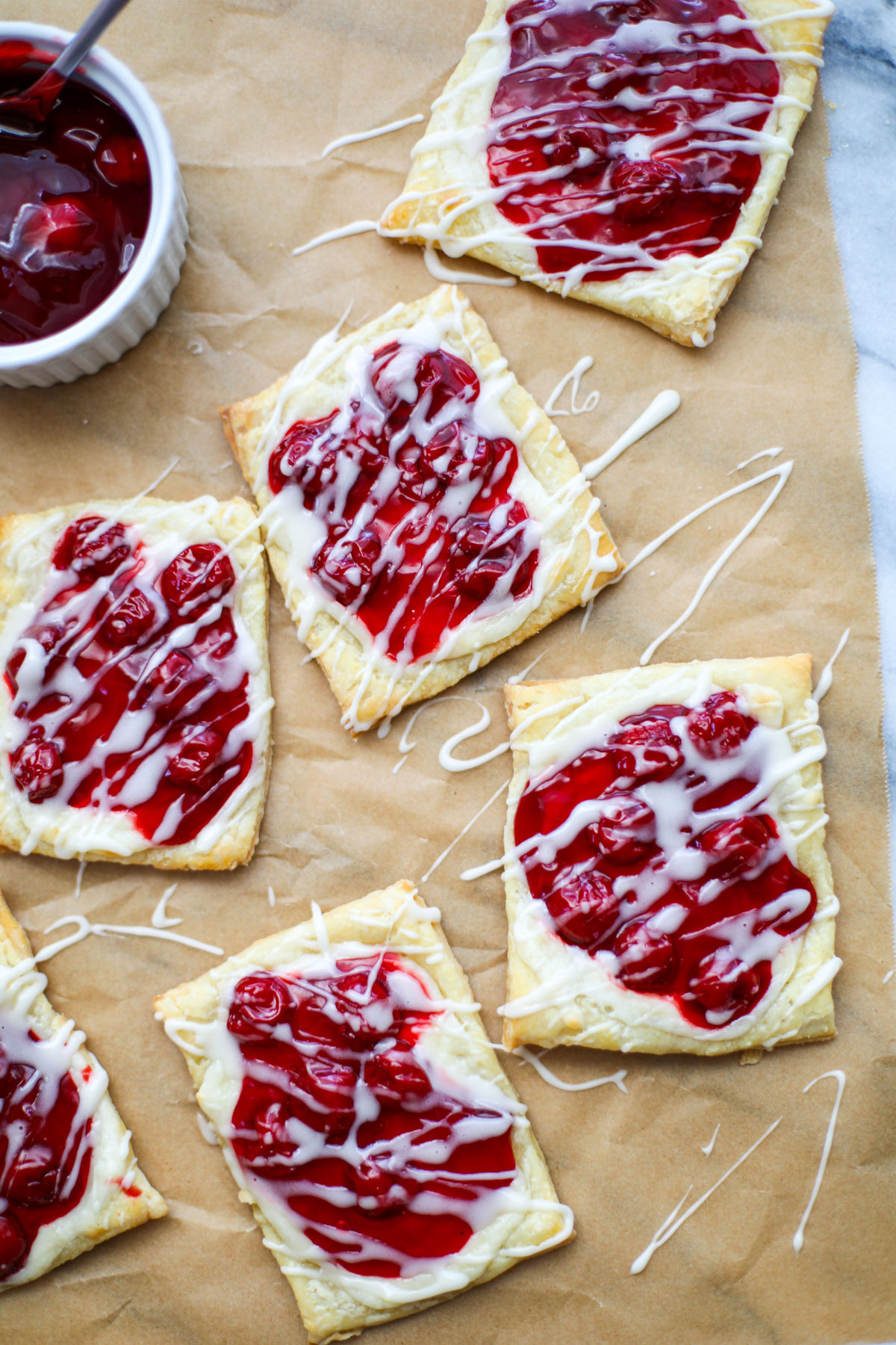Brown parchment paper topped with square cherry cheese danishes drizzled with a white glaze with a bowl of cherry pie filling in the background.