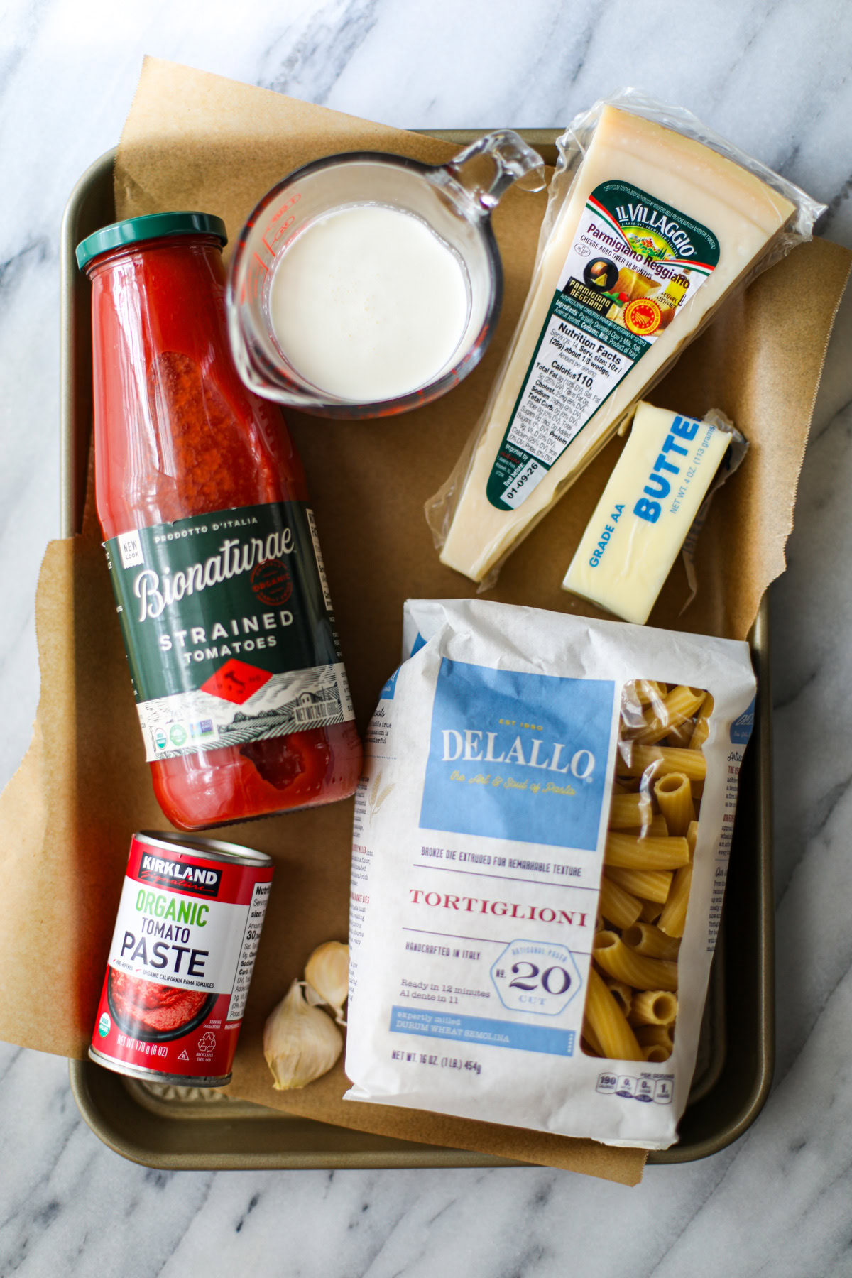 A sheet tray with ingredients to make vodka sauce including a jar of passatta, a glass measuring cup full of heavy cream, a piece of parmigiano reggiano, a blue and white bag of short cut pasta, a can of tomato paste, and half a stick of butter.
