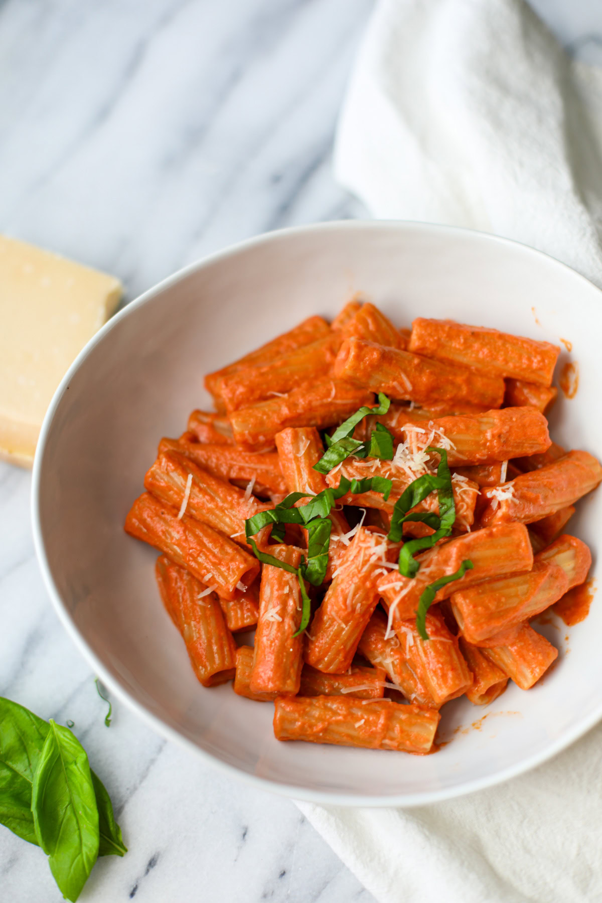 A white shallow bowl filled with vodka sauce pasta topped with cheese and chopped basil with whole basil and a block of parmigiano reggiano in the background.