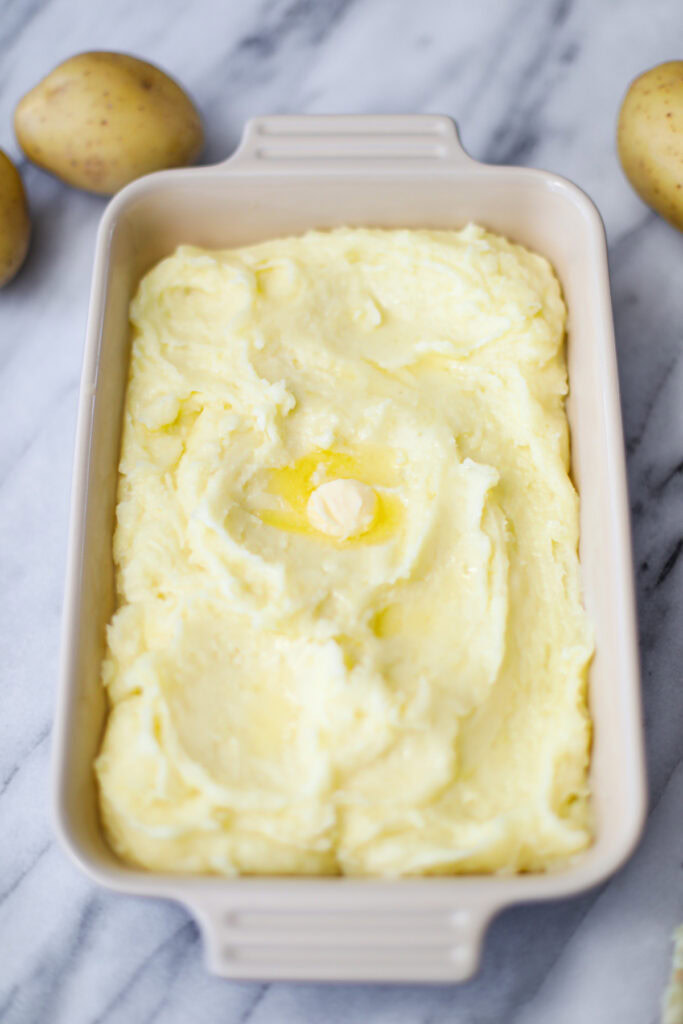 A serving dish of mashed potatoes with a nob of melted butter and potatoes surrounding the dish in the background.