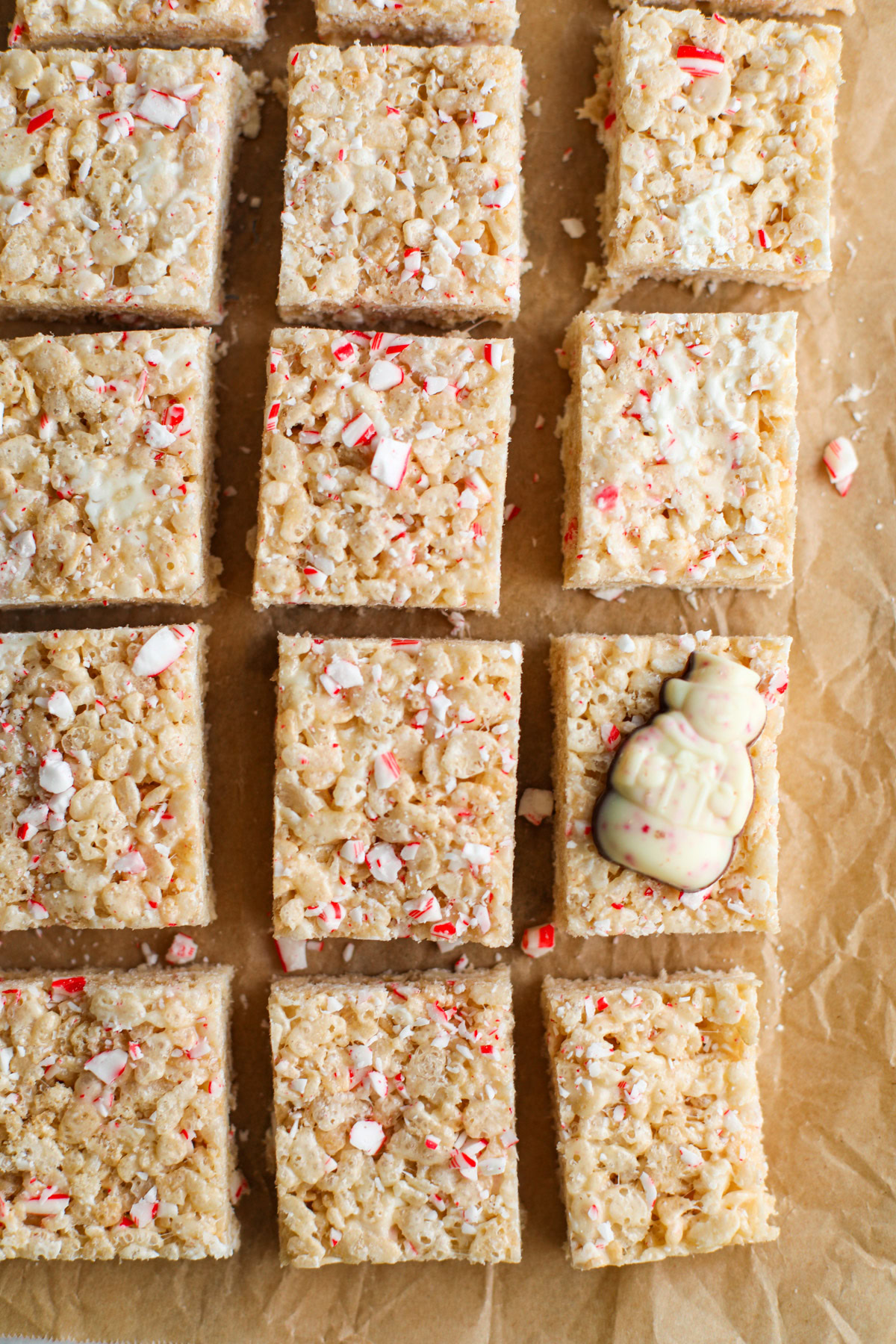 Rows of Rice Krispie treats on parchment paper topped with red and white pieces of candy cane and a peppermint bark shaped snowman candy.