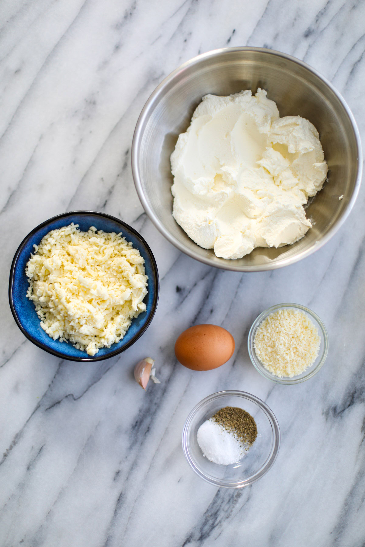 A bowl of ricotta cheese, a blue bowl of shredded mozzarella cheese, a brown egg, a large unpeeled clove of garlic, grated parmigiano and salt and black pepper in a glass bowl.