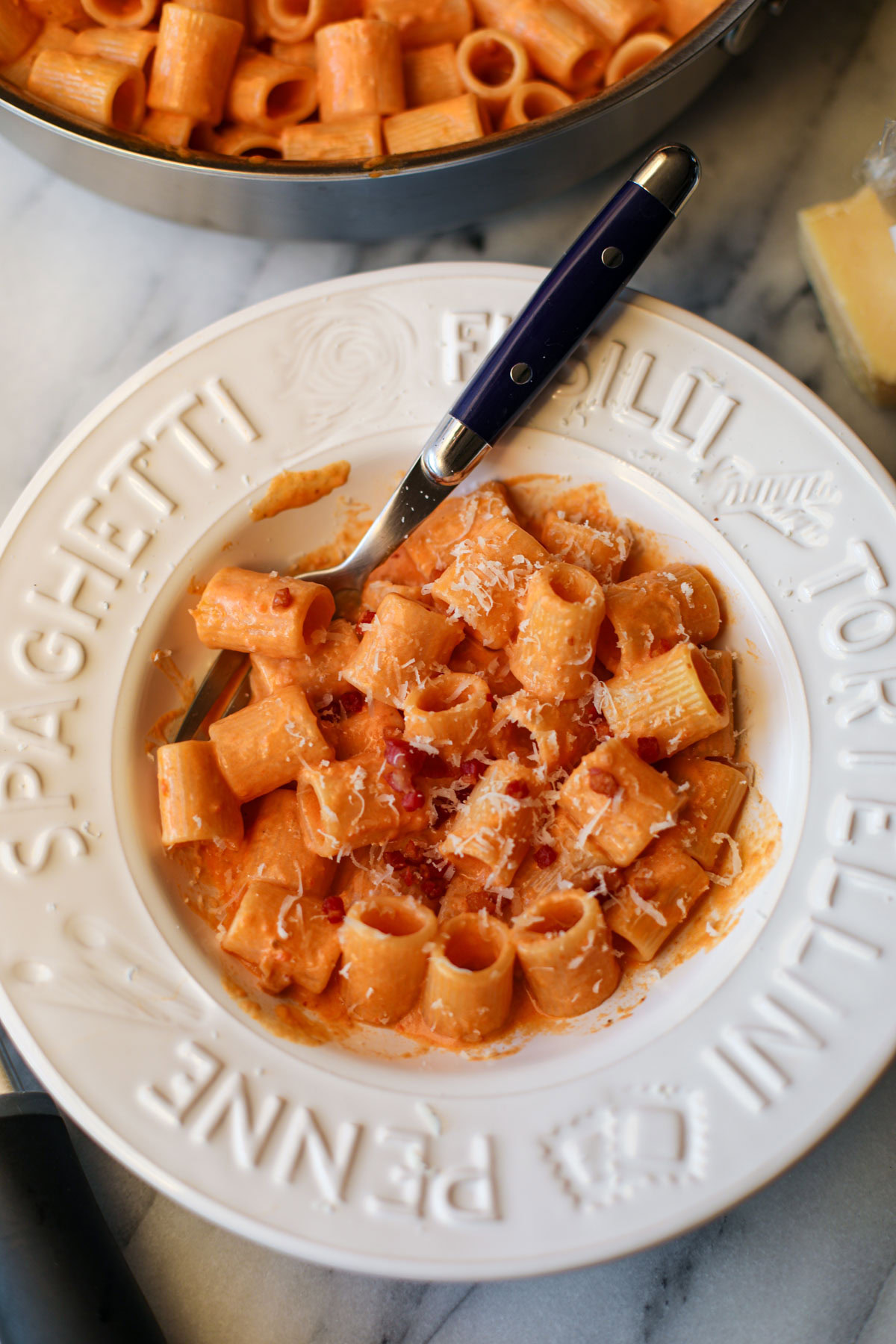 A white pasta bowl filled with blush sauce mezzi rigatoni topped with golden pancetta pieces and a blue handled fork.