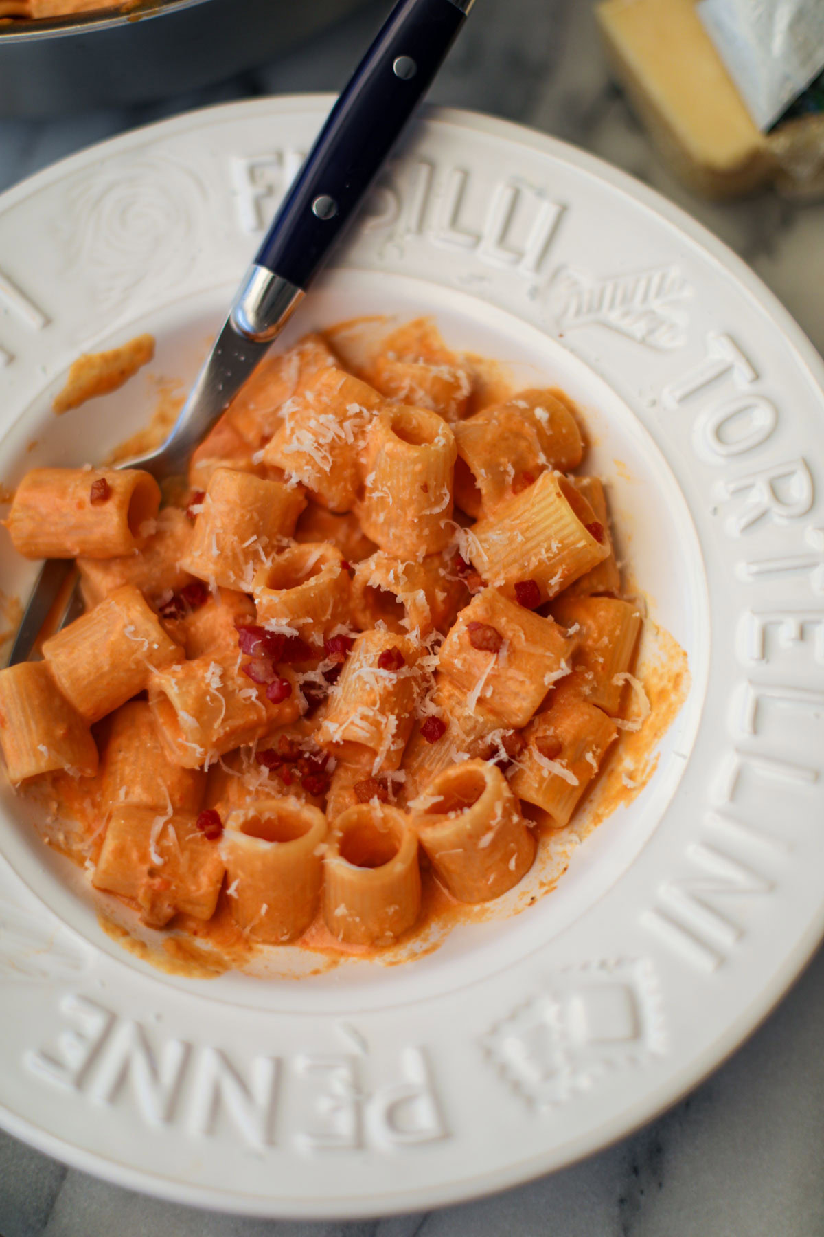 A white pasta bowl filled with blush sauce mezzi rigatoni topped with golden pancetta pieces and a blue handled fork.