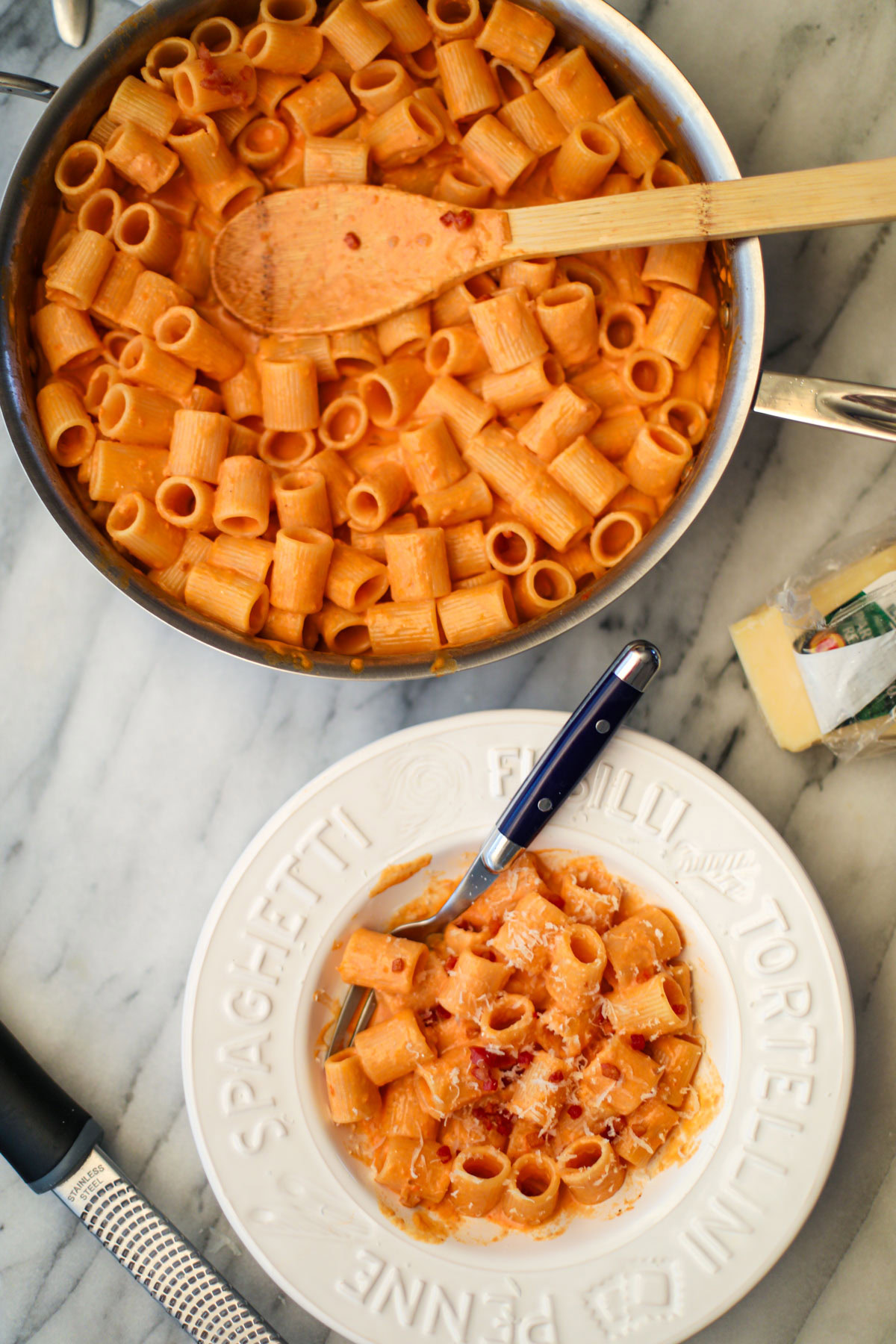A white pasta bowl filled with blush sauce mezzi rigatoni topped with golden pancetta pieces and a blue handled fork with a silver skillet of pasta in the background.