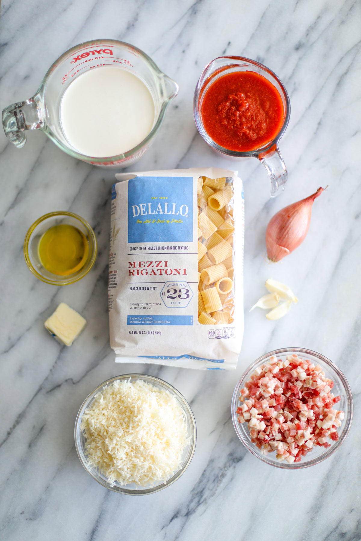 A measuring cup filled with cream and marinara sauce, a blue and white package of pasta, a nob of butter, a whole shallot, 3 garlic gloves, and glass bowls filled with olive oil, grated parmigiano, and diced pancetta.