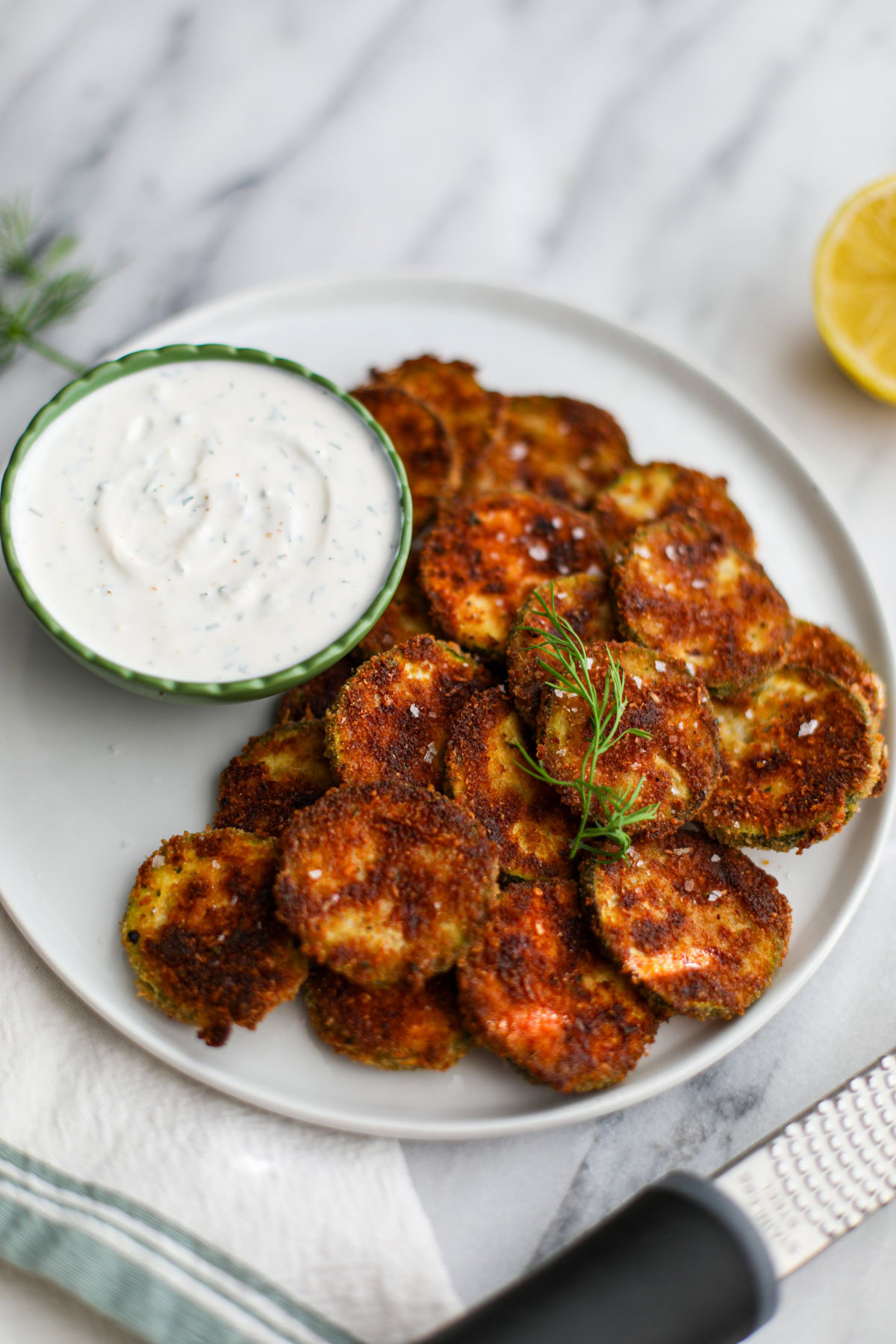 Oven baked zucchini coins on a white plate with a ruffled green bowl filled with dill dipping sauce with a lemon in the background.
