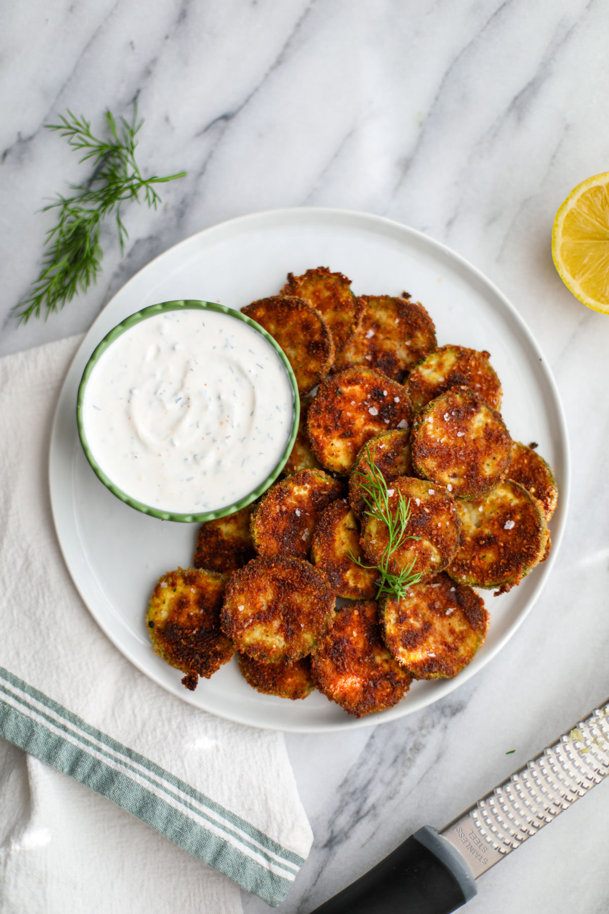 Oven baked zucchini coins on a white plate with a ruffled green bowl filled with dill dipping sauce with a microplane grater and half a lemon.