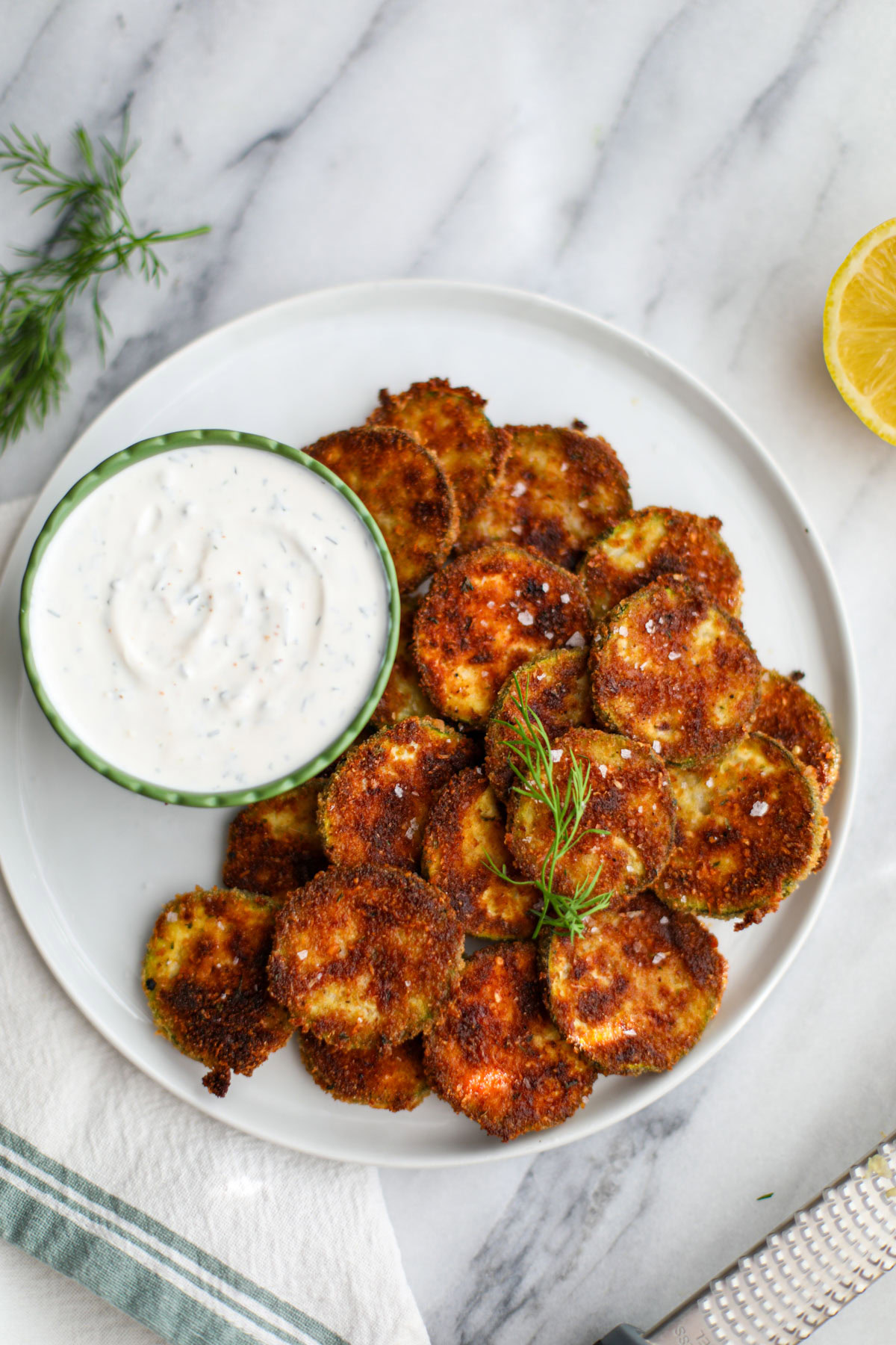 Oven baked zucchini coins on a white plate with a ruffled green bowl filled with dill dipping sauce.