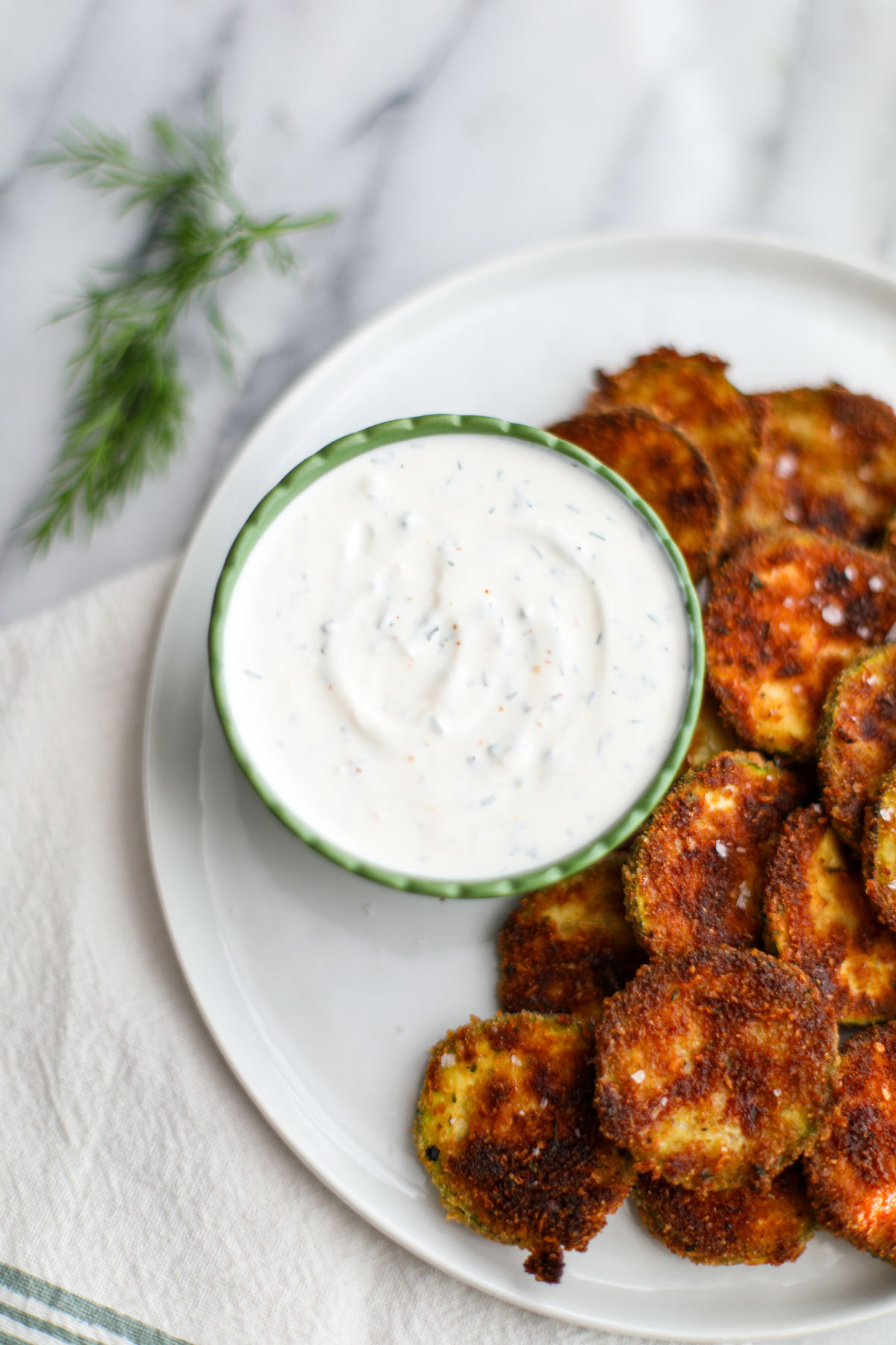 A small ruffled green sauce bowl filled with white dill sauce next to zucchini coins.