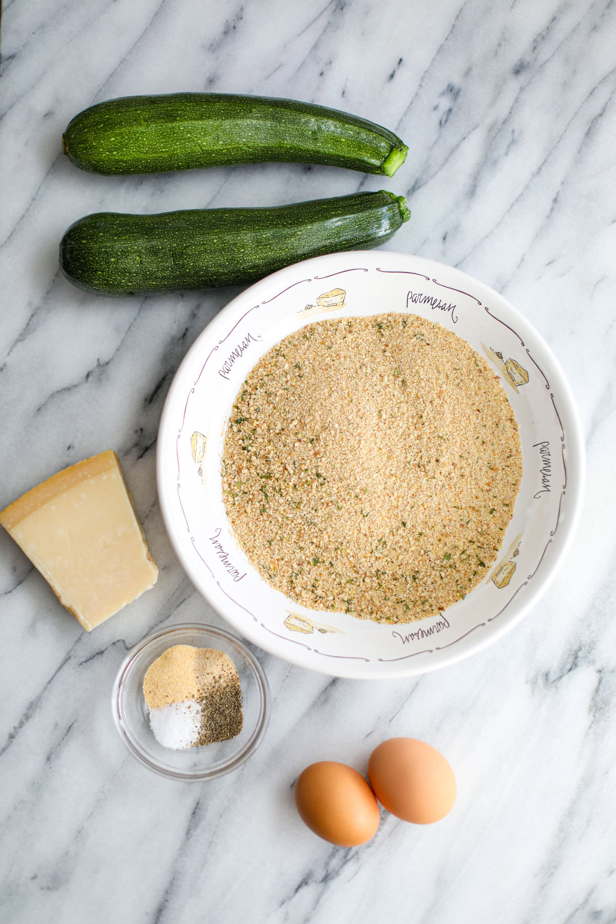 Two zucchinis, a white bowl of bread crumbs, a hunk of parmigiano reggiano, a clear bowl of seasonings, and 2 eggs.