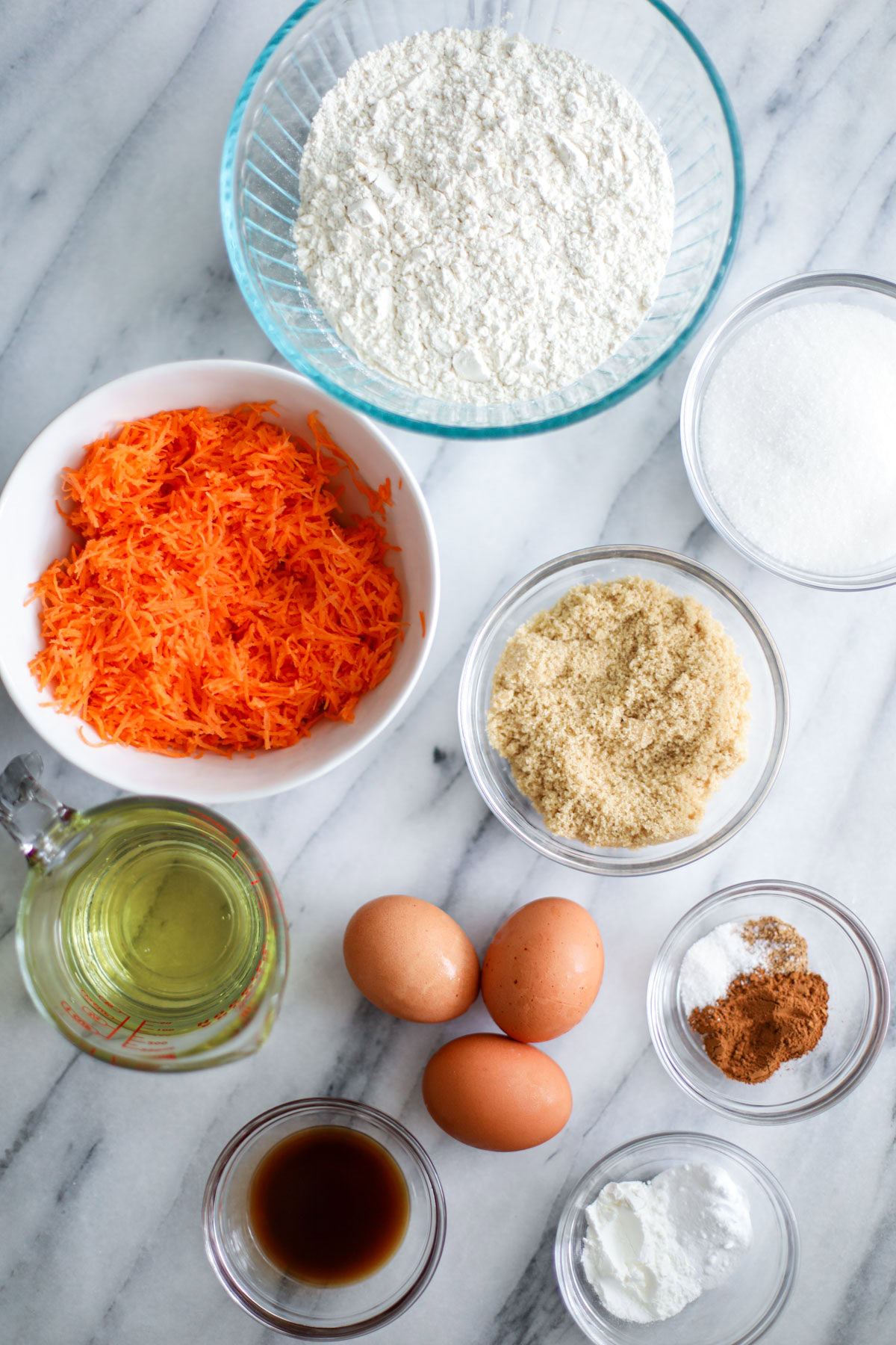 Glass bowls of ingredients including flour, white sugar, brown sugar, grated carrots, cinnamon and salt, oil, vanilla extract, baking powders, and 3 eggs in the center.