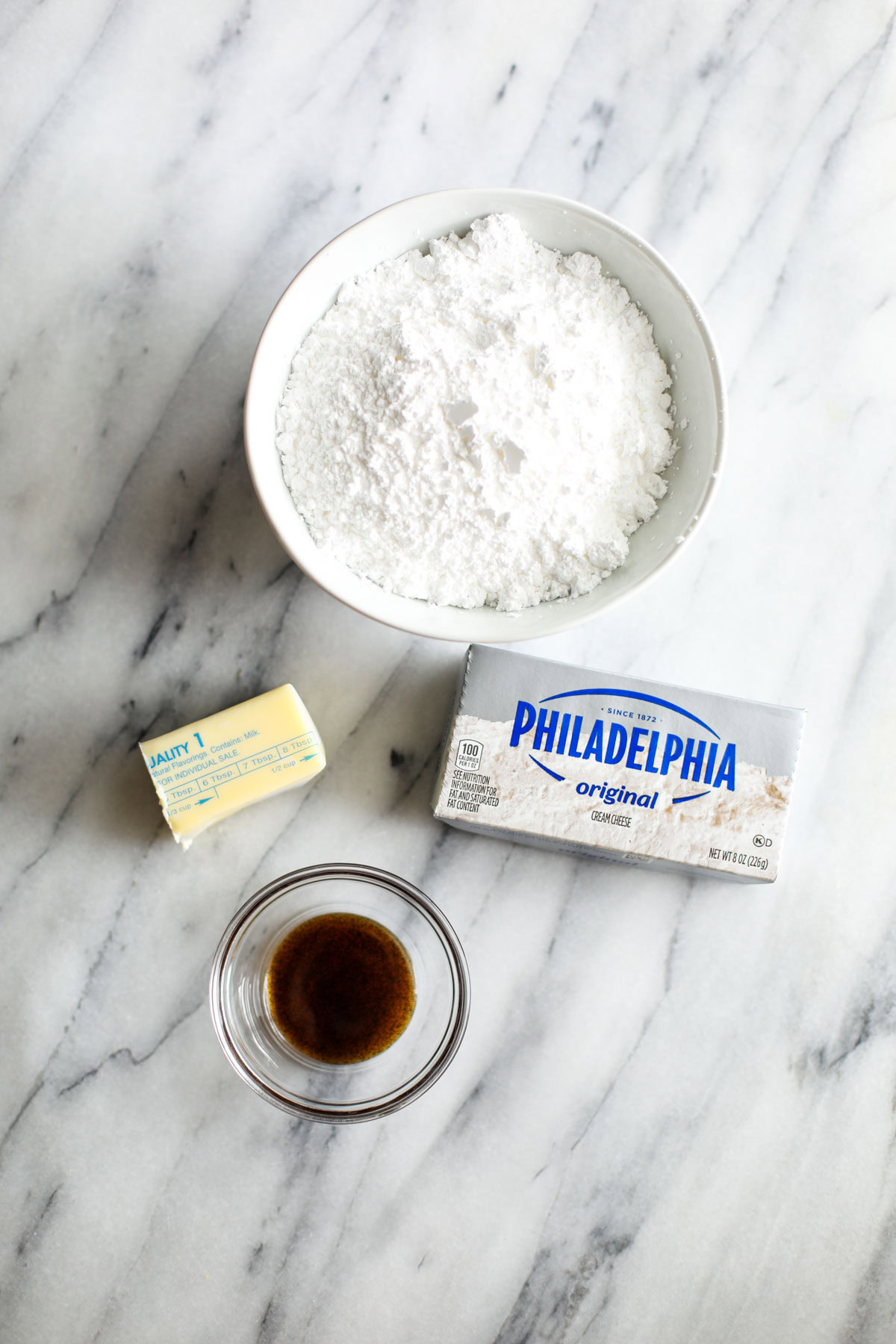 A bowl of powdered sugar, a block of wrapped Philadelphia cream cheese, half a stick of wrapped butter, and a glass bowl of vanilla bean paste.