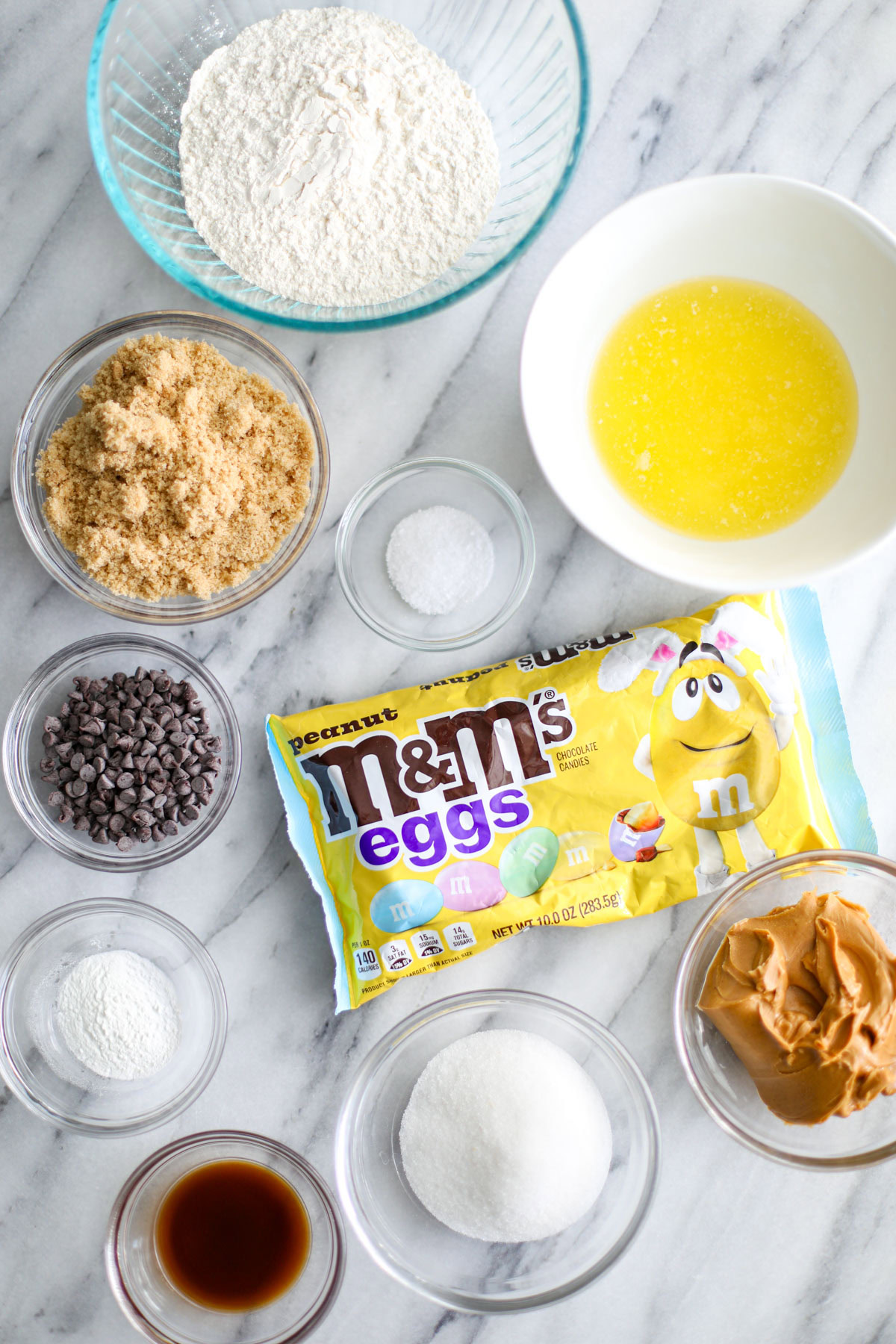A glass bowl of flour, light brown sugar, mini chocolate chips, salt, vanilla, baking powder, white sugar, and peanut butter with a white bowl of melted butter and a yellow bag of peanut M&M's eggs.