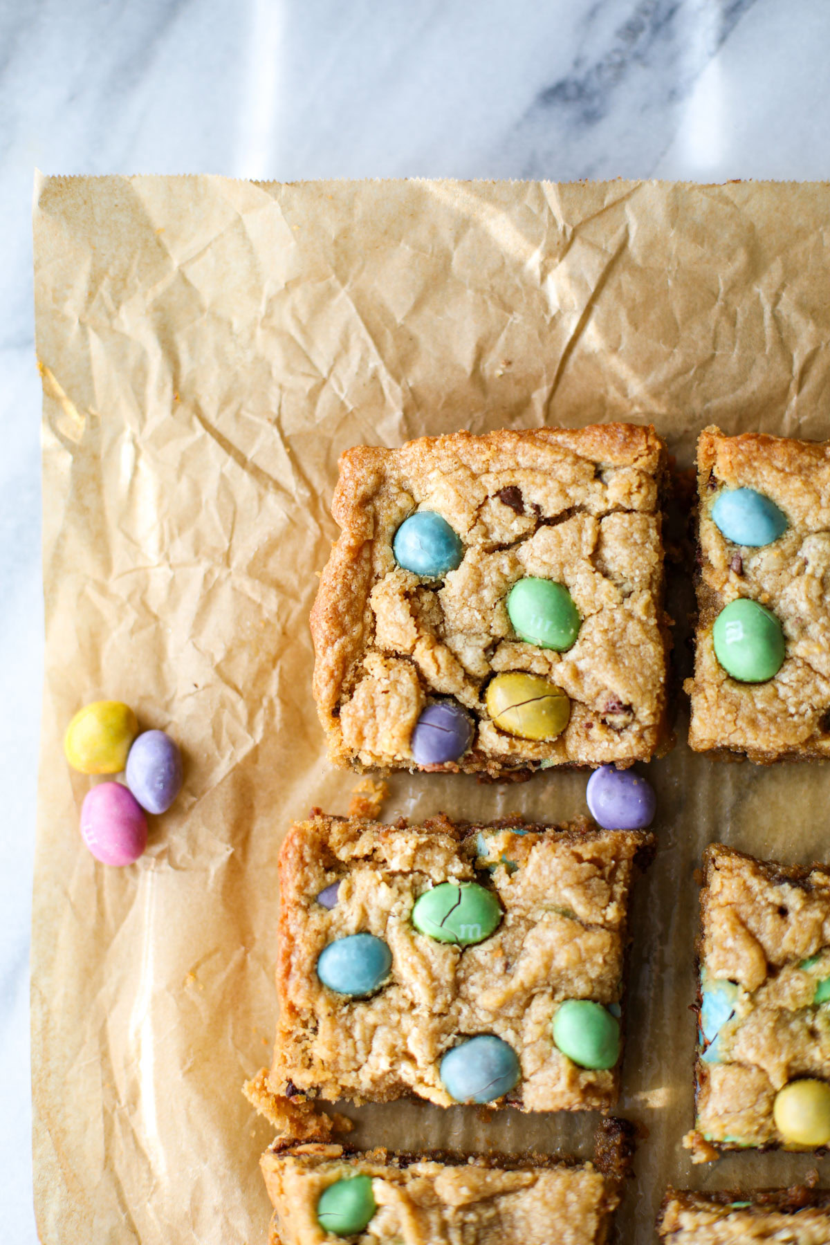 Peanut M&M Easter Egg Blondies studded with green, yellow, pink, and purple m&ms on brown crinkled parchment paper lined up next to other blondies. 