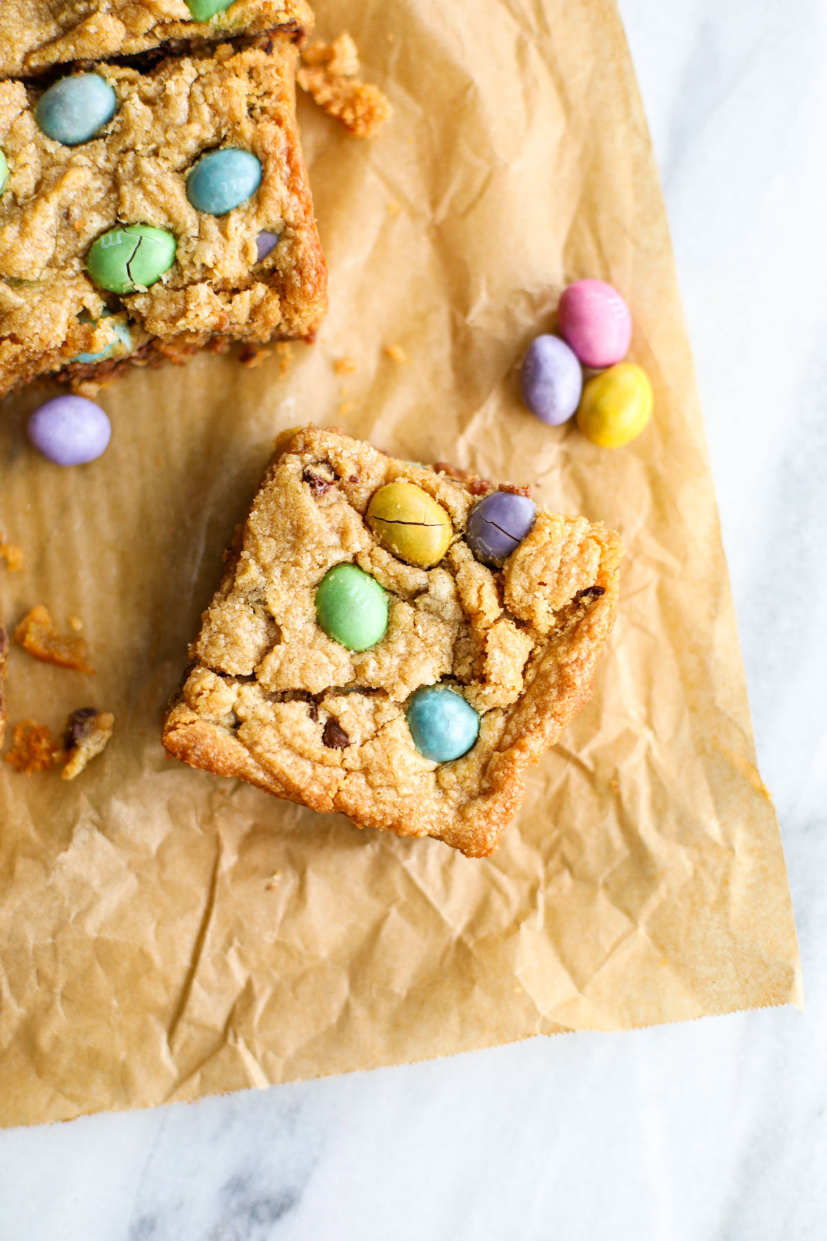 A Peanut M&M Easter Egg Blondie studded with green, yellow, pink, and purple m&ms on brown crinkled parchment paper.