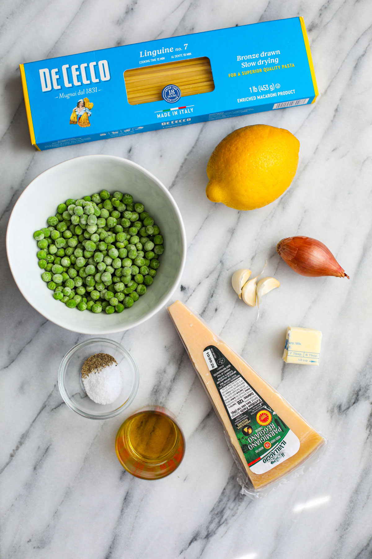 A carton of de cecco pasta, a whole lemon, a white bowl of peas, a shallow, 3 cloves of garlic, a wedge of parmigiano, a glass cup of olive oil, a small bowl of salt, and 2 tablespoons of butter. 
