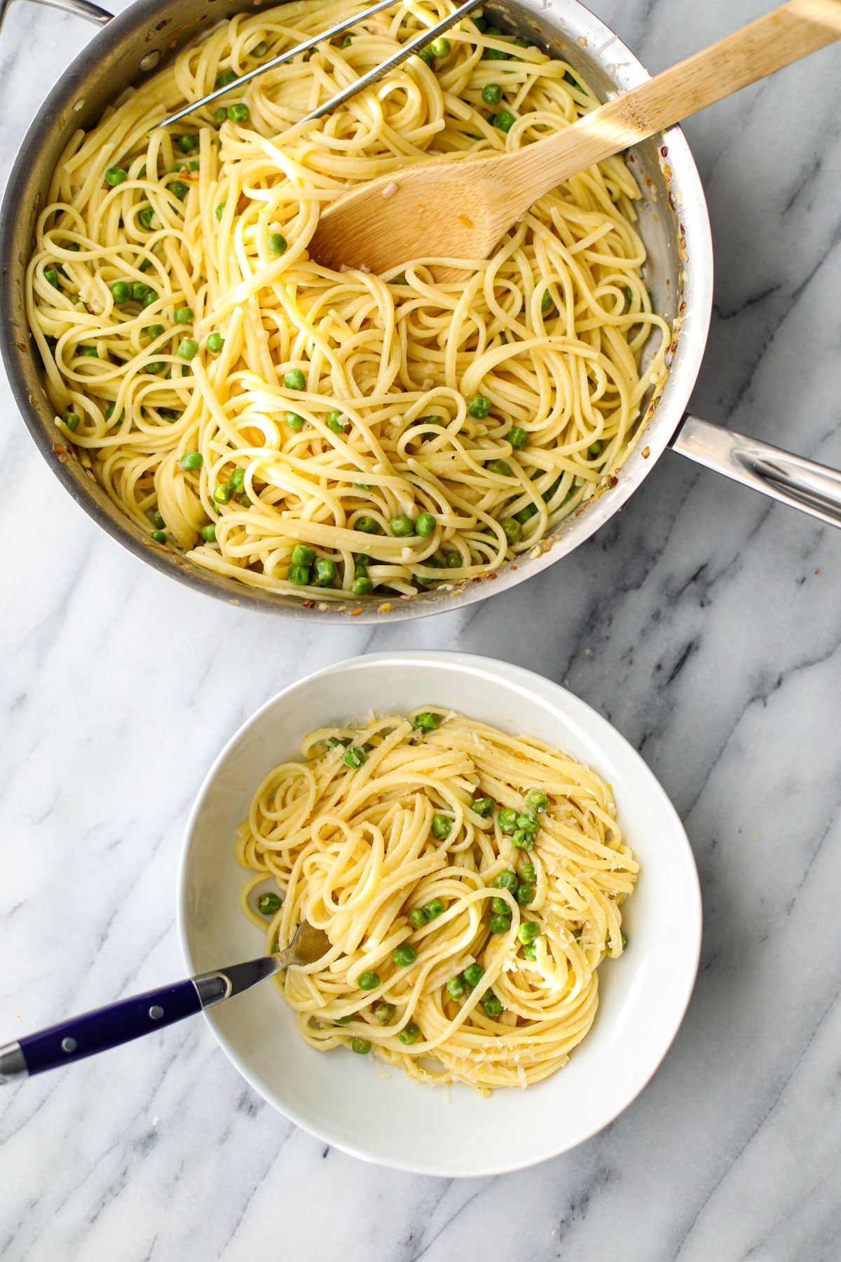 Lemon pasta with peas in a stainless steel pan with a wooden spoon and metal tongs with a white bowl off to the side. 