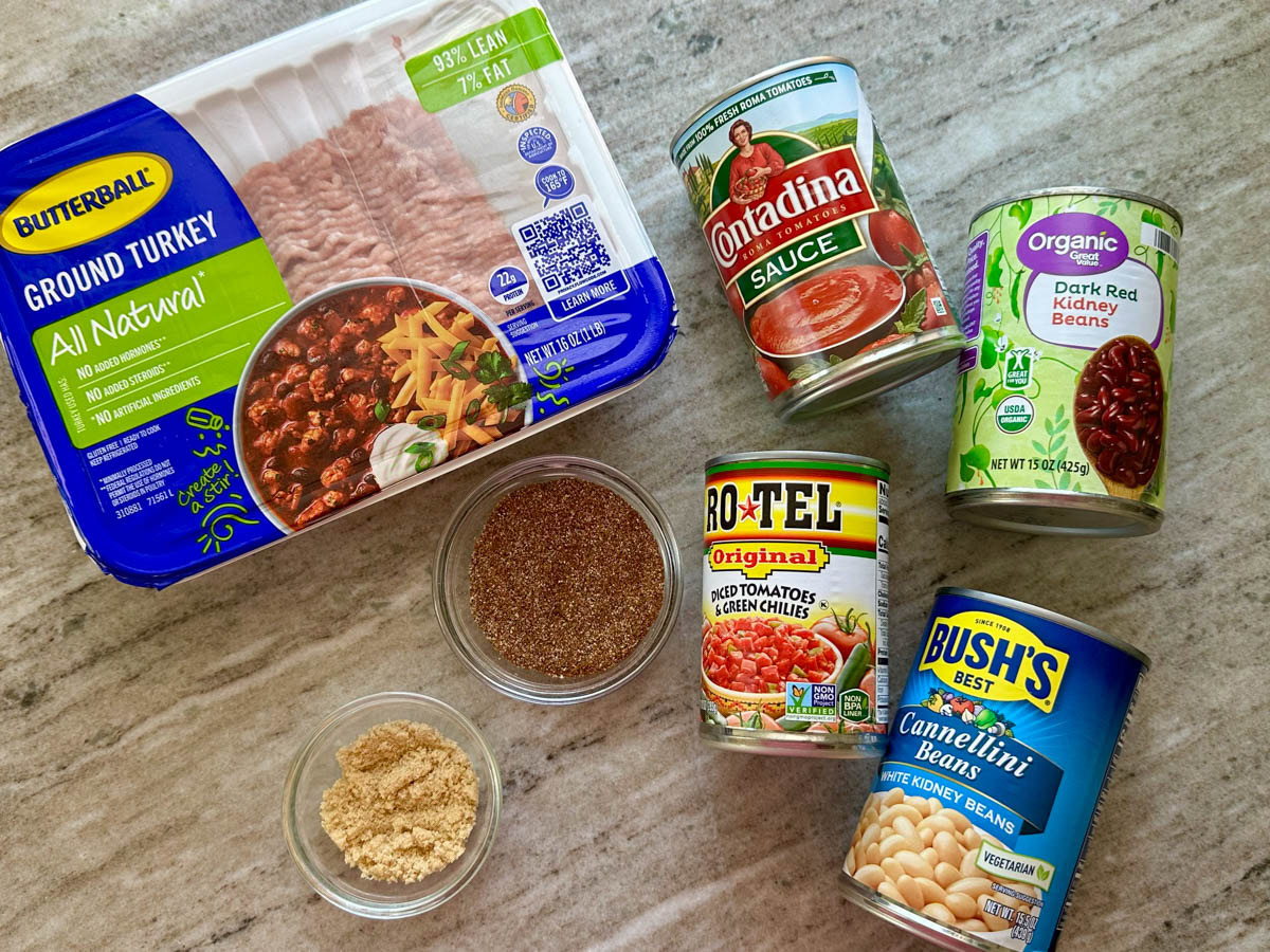 Ingredients on the table top to make chili including ground turkey, cans of tomato sauce, red kidney beans, white kidney beans, diced tomatoes and green chilies and chili seasonings.