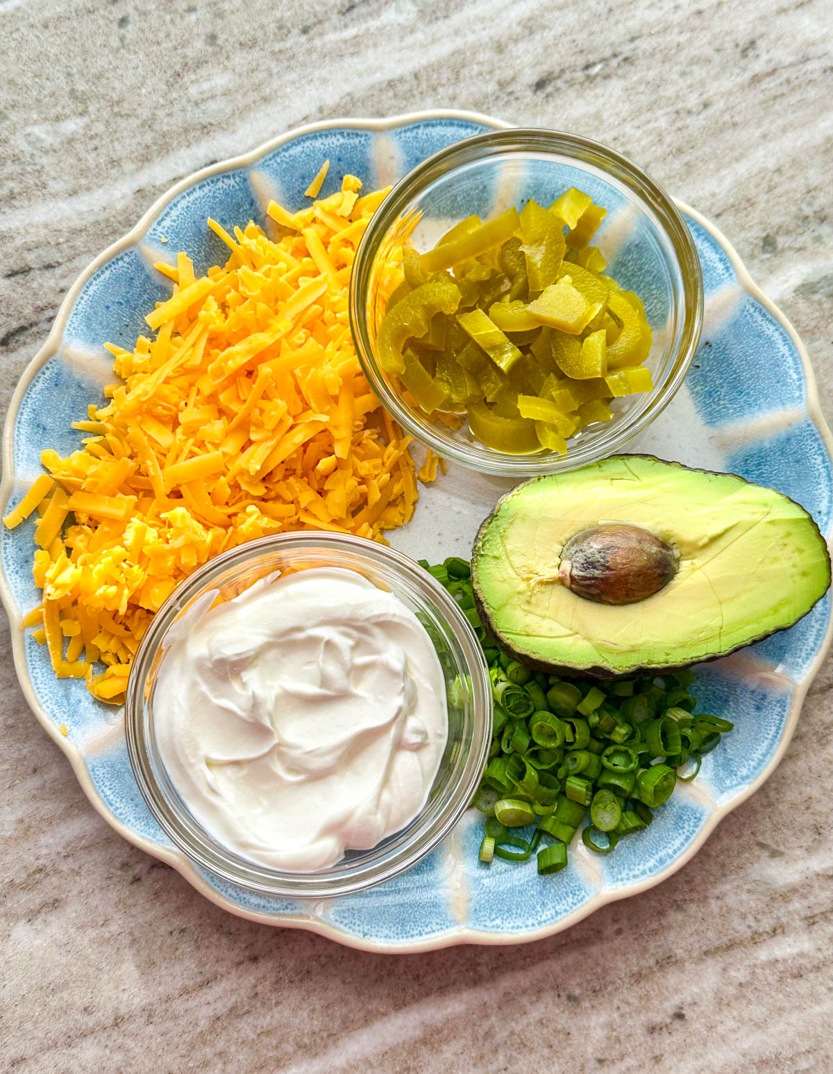 A blue plate filled with chili toppings including shredded sharp cheddar cheese, sour cream, jalapeños, avocado, and green onion.