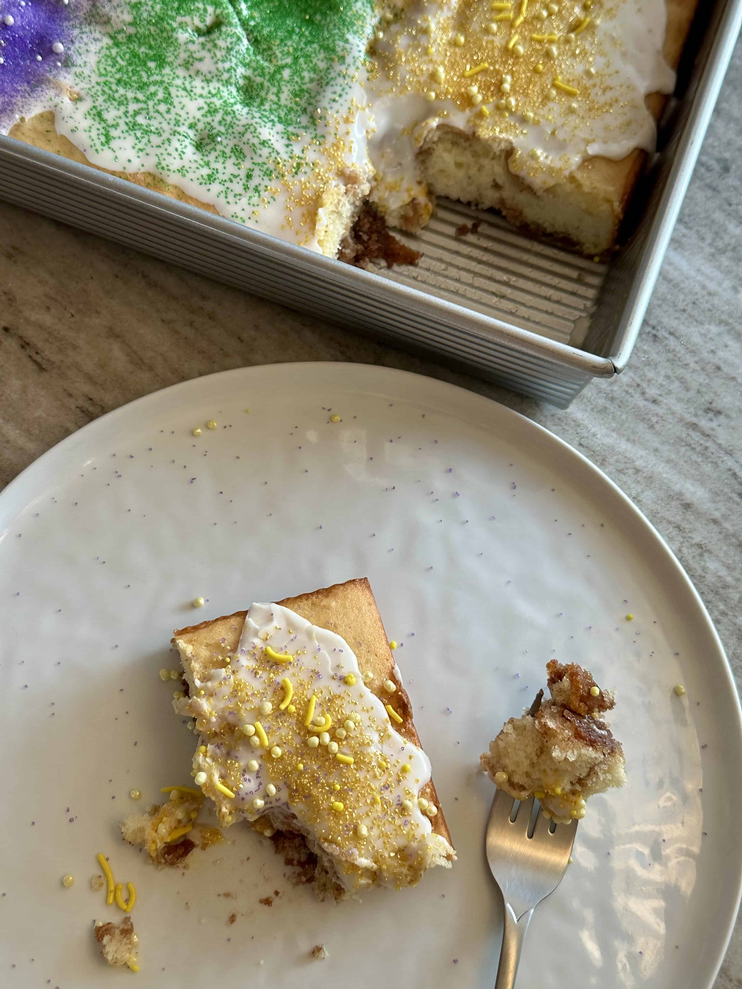 A white plate with a slice of king cake with the cake pan in the background with the king cake inspired sheet cake.
