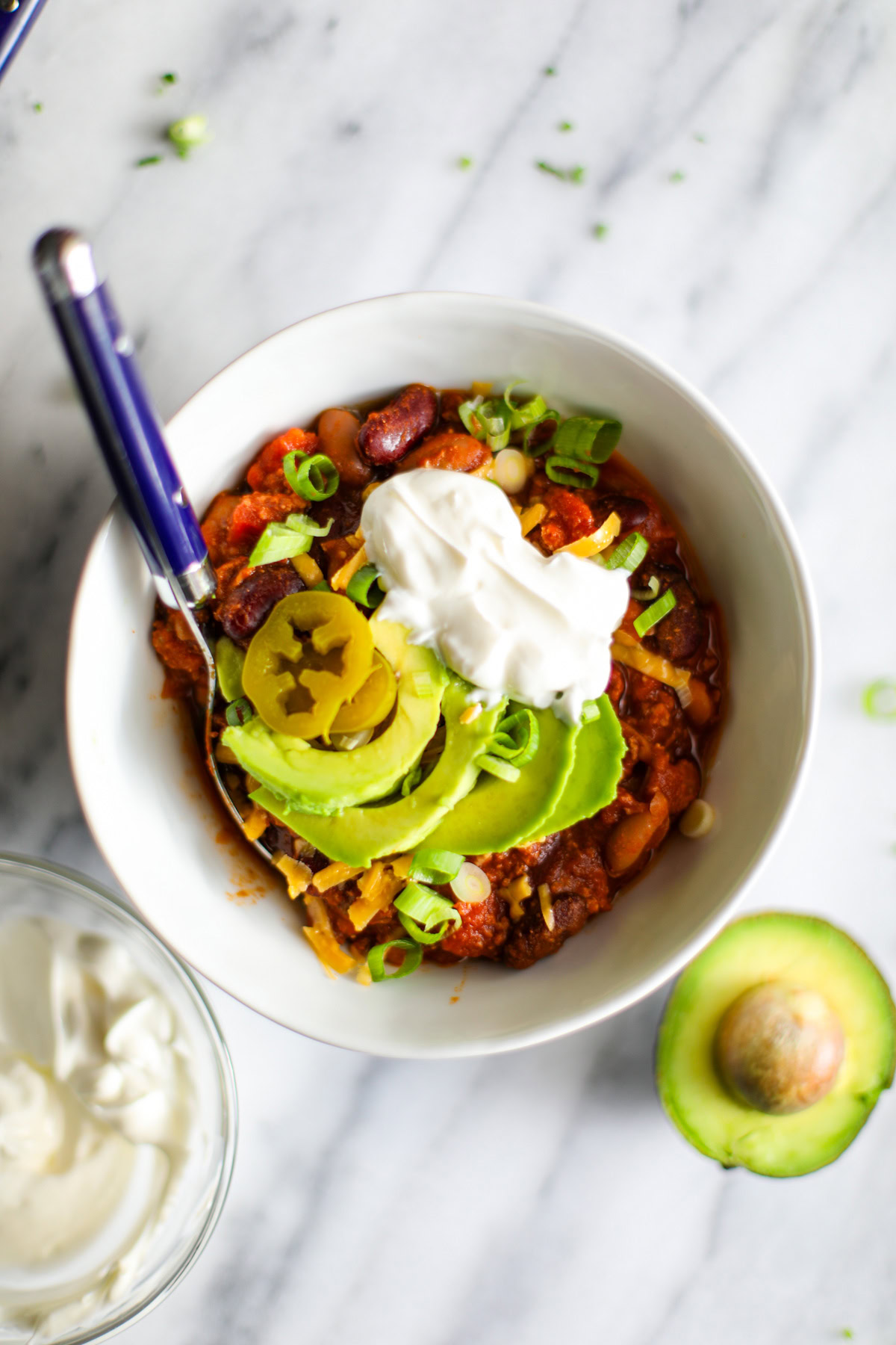 A white bowl filled with turkey chili and topped with cheddar cheese, avocado, sour cream, and green onions. In the background is a blue plate with the chili toppings and hot sauce.