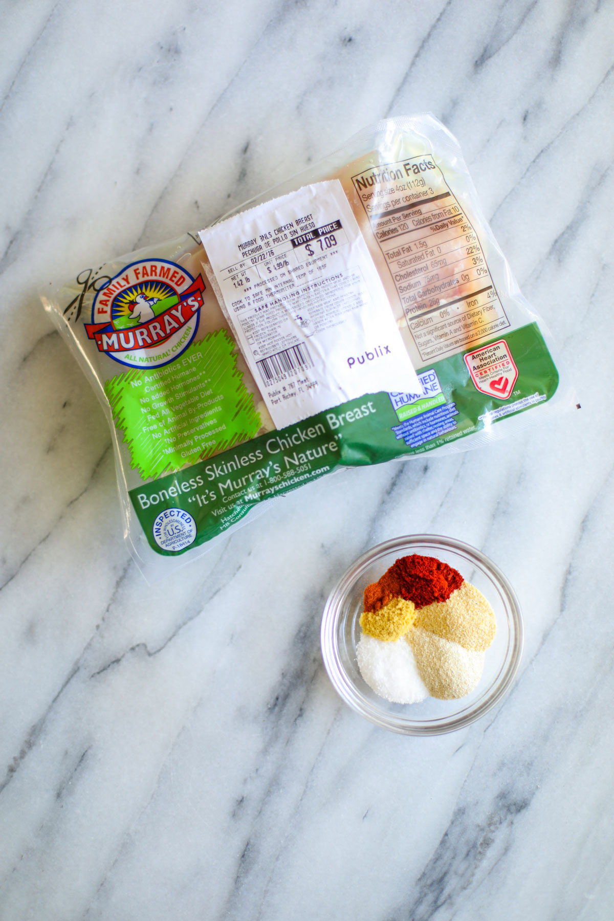 A green store package of chicken breasts and a clear bowl of yellow, red, and white seasonings.