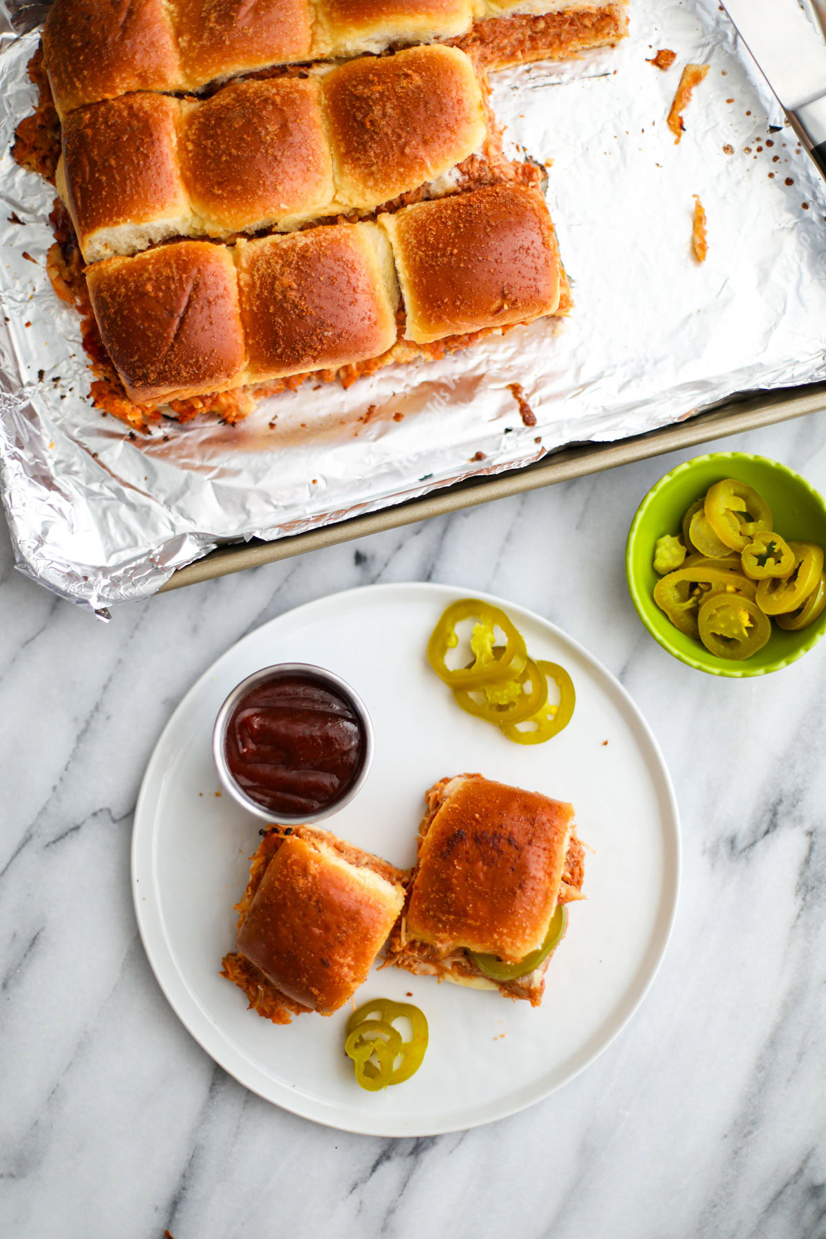 Two BBQ chicken sliders on a white plate with pieces of jalapeños and a stainless steel cup of BBQ sauce next to a sheet tray of sliders.