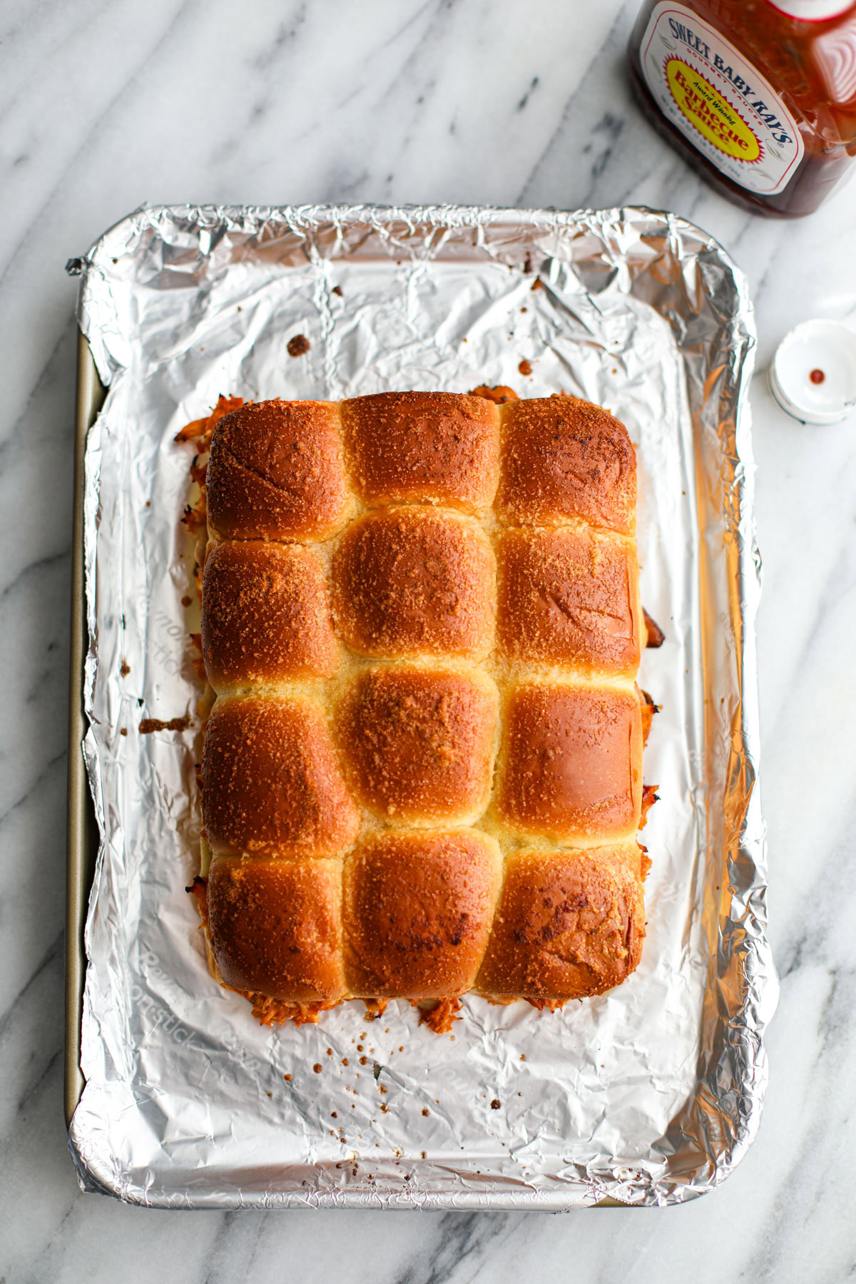 Rows of uncut BBQ Chicken Sliders with golden tops and melty cheese on a foil lined pan with a bottle of barbecue sauce.