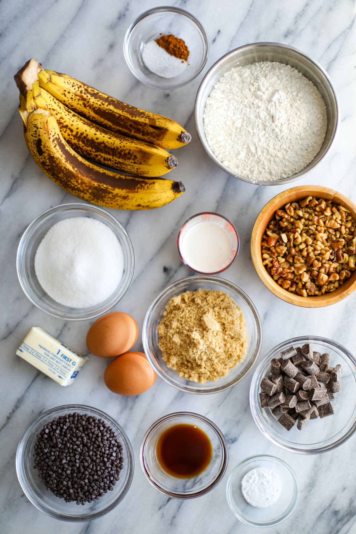Three very ripe bananas, a bowl of flour, sugar, salt and cinnamon, chopped walnuts, chocolate chips, vanilla, a stick of butter, and 2 eggs on a marble counter.