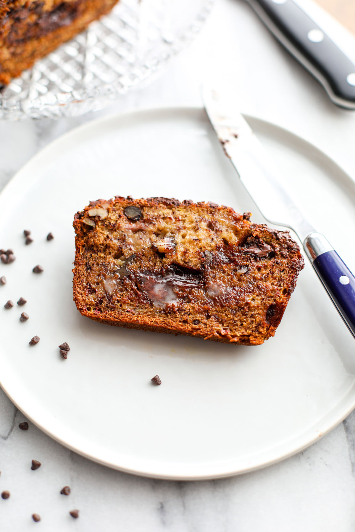 A slice of banana bread on a white plate with a blue knife and mini chocolate chips.