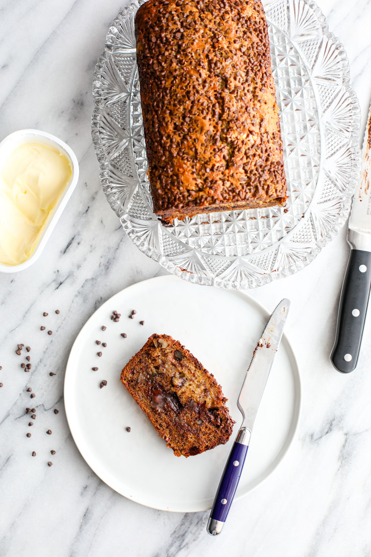 A loaf of chocolate chip walnut banana bread on a clear patter with a white plate with a slice of bread and a tub of butter.