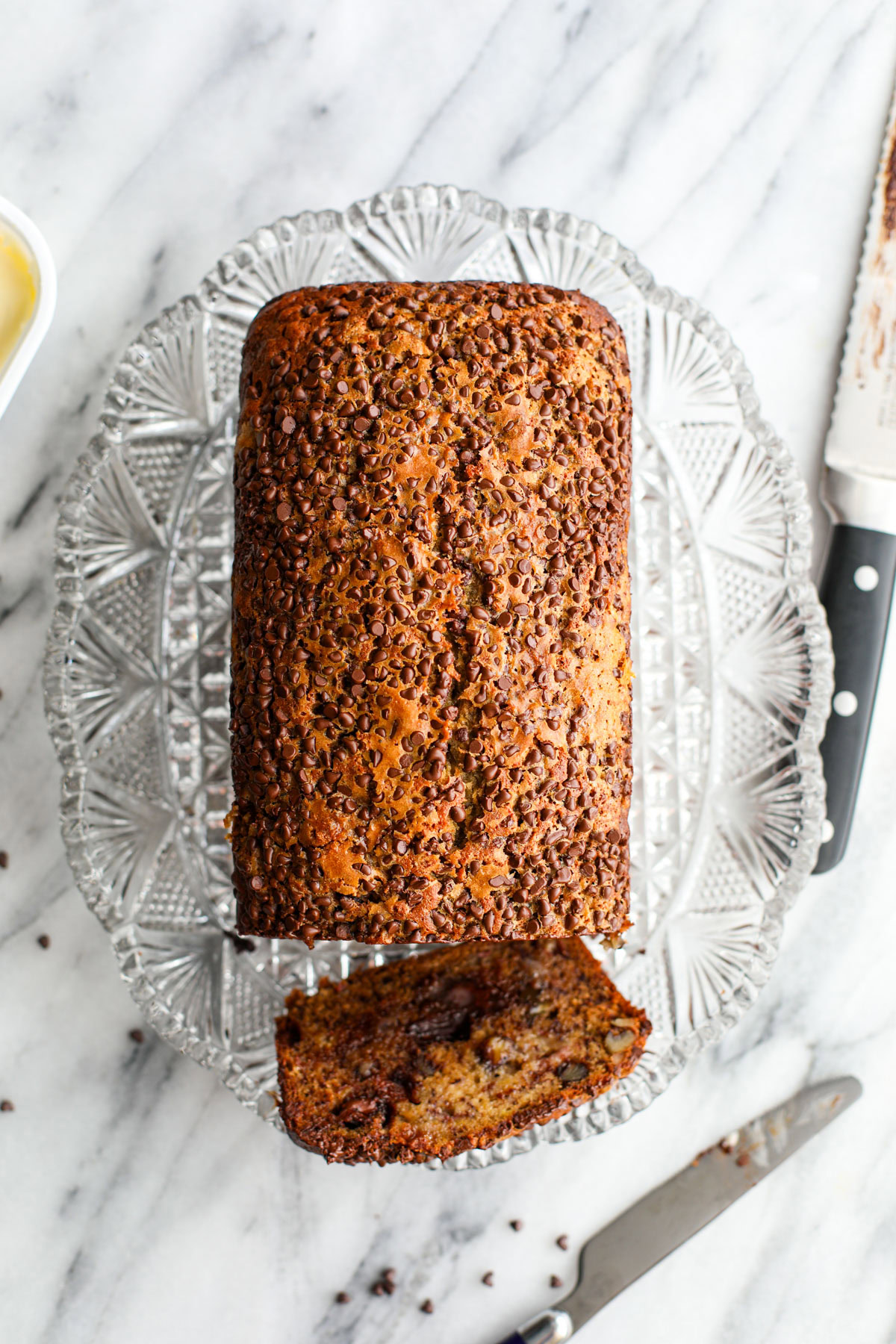 A loaf of chocolate chip topped banana bread on a glass platter with a slice in front of it.