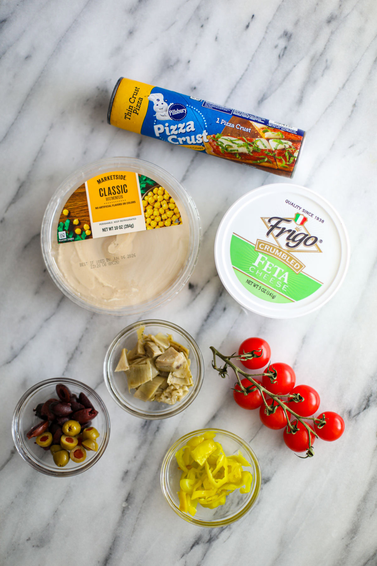 Clear bowls filled with peppers, marinated artichoke hearts, feta, chopped red onion, cherry tomatoes, green and Kalamata olives and a dish of hummus with a pillsbury canned pizza crust.