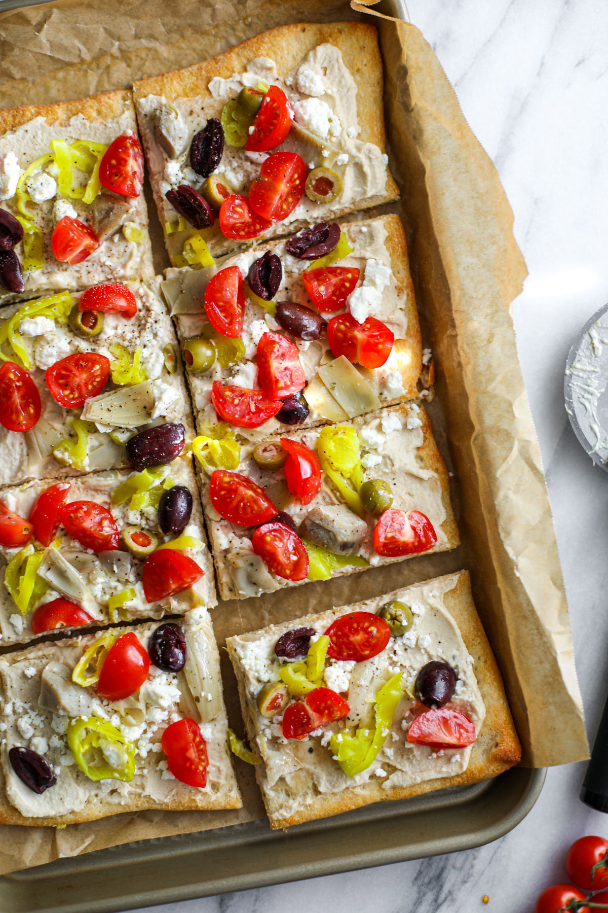 A mediterranean pizza sitting on a marble cutting board topped with hummus, feta, cherry tomatoes, olives, artichoke hearts, and red onion with loose olives in the background.