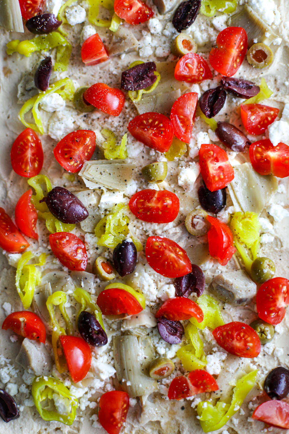 A close up of the toppings on the Mediterranean hummus pizza including olives, tomatoes, feta, pepperoncini, and artichoke hearts. 
